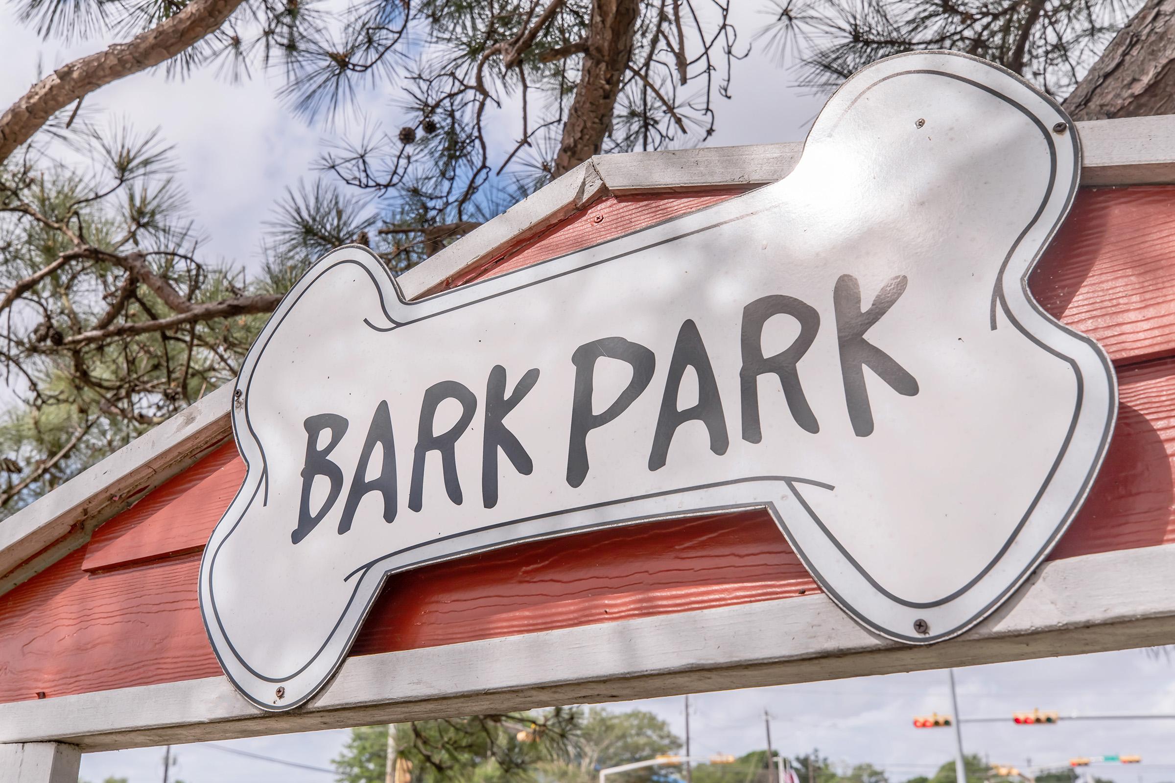 A sign shaped like a dog bone displaying the words "BARK PARK" in bold lettering, hanging from a wooden structure. The background features trees and a cloudy sky, indicating an outdoor dog park setting.