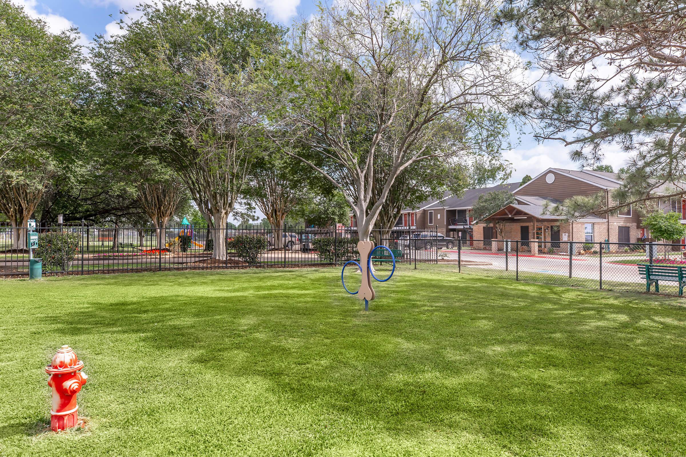 A sunny park scene featuring a well-maintained green lawn, a decorative playground structure in the background, and a fenced area. In the foreground, there is a red fire hydrant and a unique sculpture with circular elements. The park is surrounded by trees, providing shade and a welcoming atmosphere.