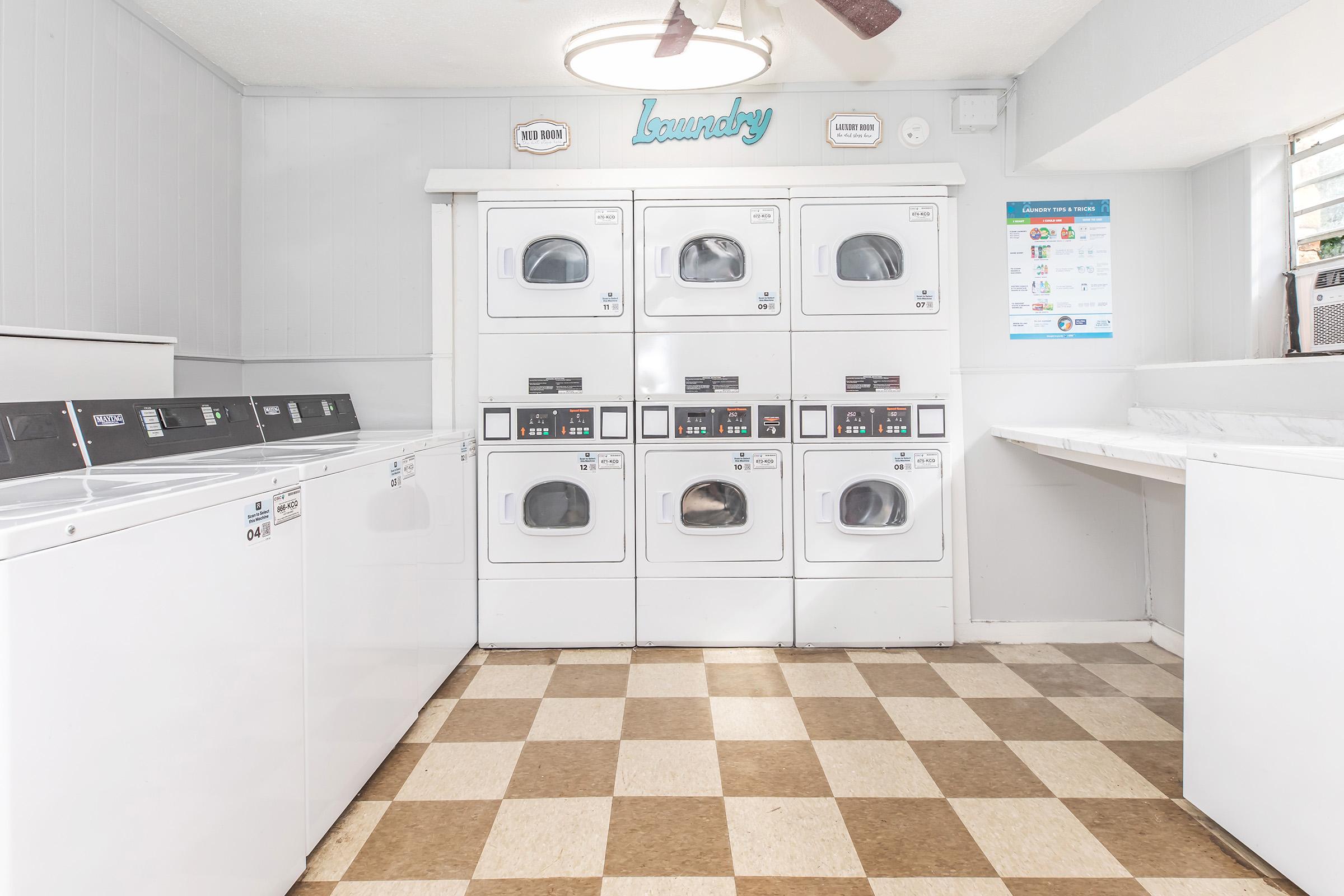 A clean laundry room featuring several white washing machines and dryers arranged in a row. The floor has a checkered pattern with beige and brown tiles. A sign labeled "Laundry" is displayed on the wall, and there is a flat surface for folding clothes beside the machines.