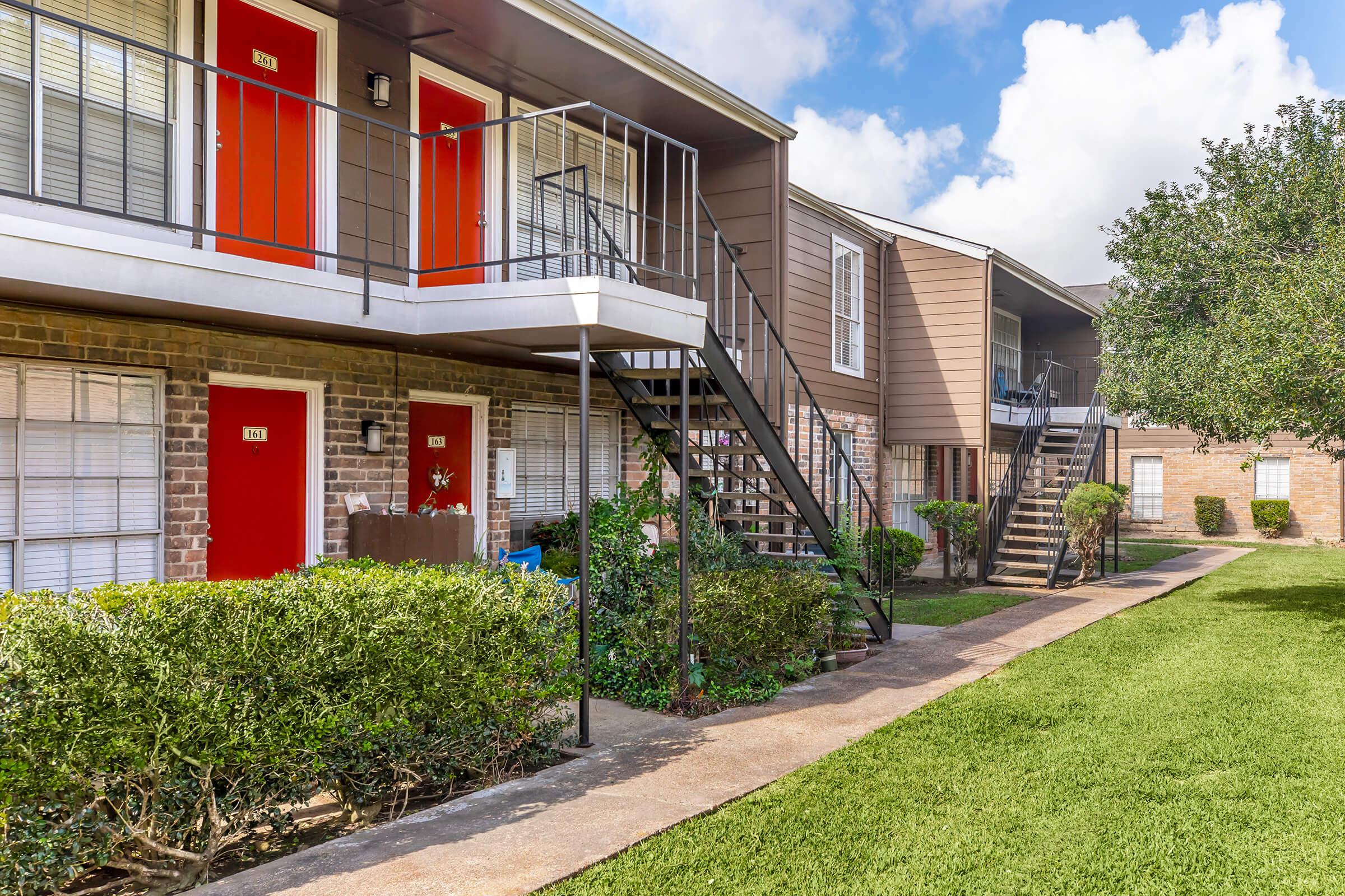 A view of an apartment complex featuring two-story buildings with red doors and metal staircases. The ground level is bordered by neatly trimmed bushes and a grassy lawn, with a clear blue sky above and some fluffy clouds. The surroundings are well-maintained, creating a welcoming atmosphere.