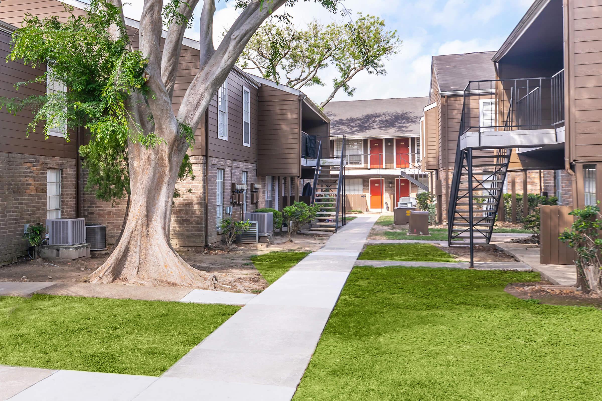 A landscaped courtyard of an apartment complex featuring a paved walkway, greenery, and brown wooden buildings. Trees and plants are scattered around the pathway, leading to entrances of the apartments. Some units have outdoor staircases and railings. The sky is partly cloudy.
