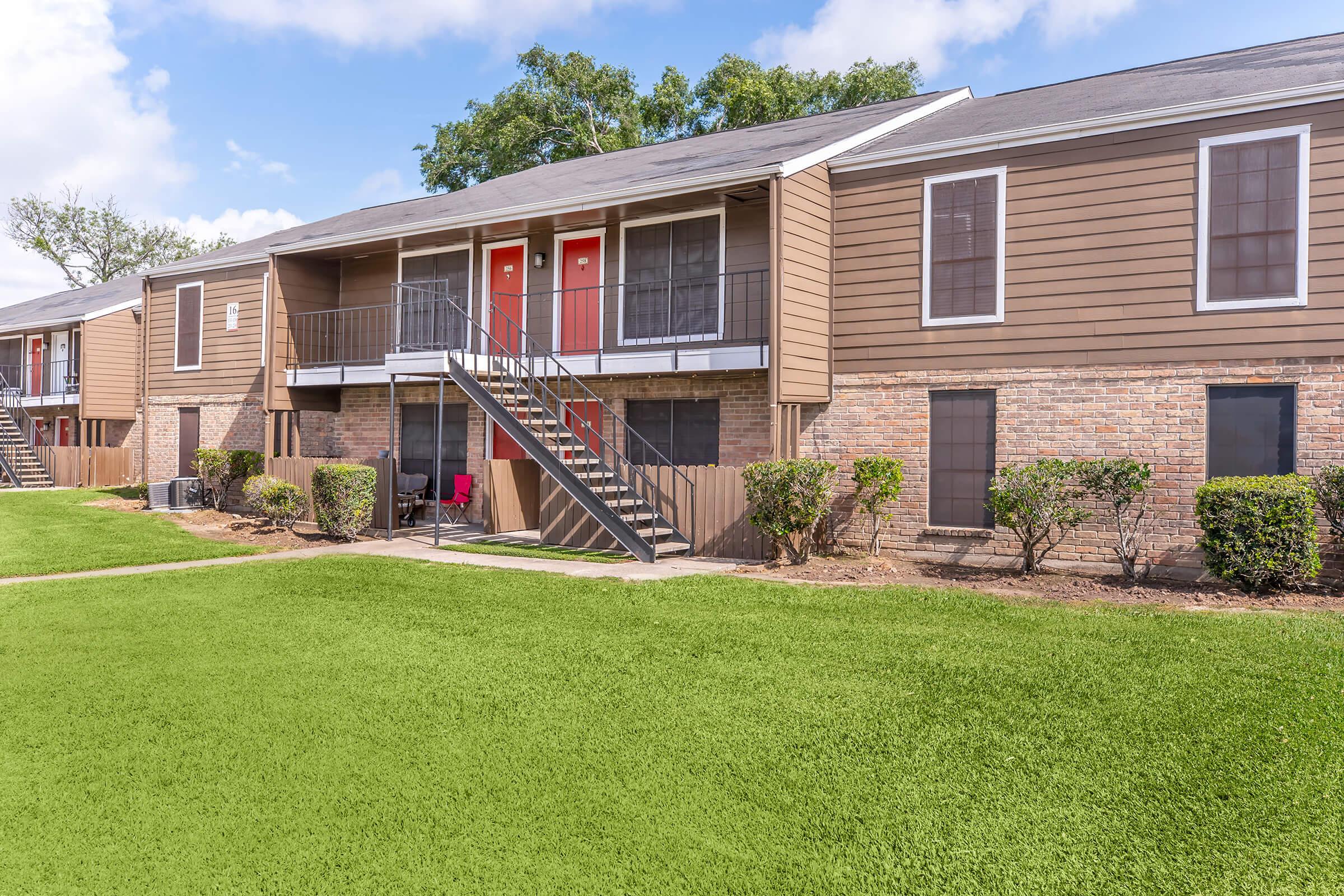 A view of a two-story apartment building with brown siding and red doors. The building features balconies and a staircase leading to the upper units. In the foreground, there is a well-maintained green lawn and small shrubs, set against a backdrop of blue skies with some clouds.
