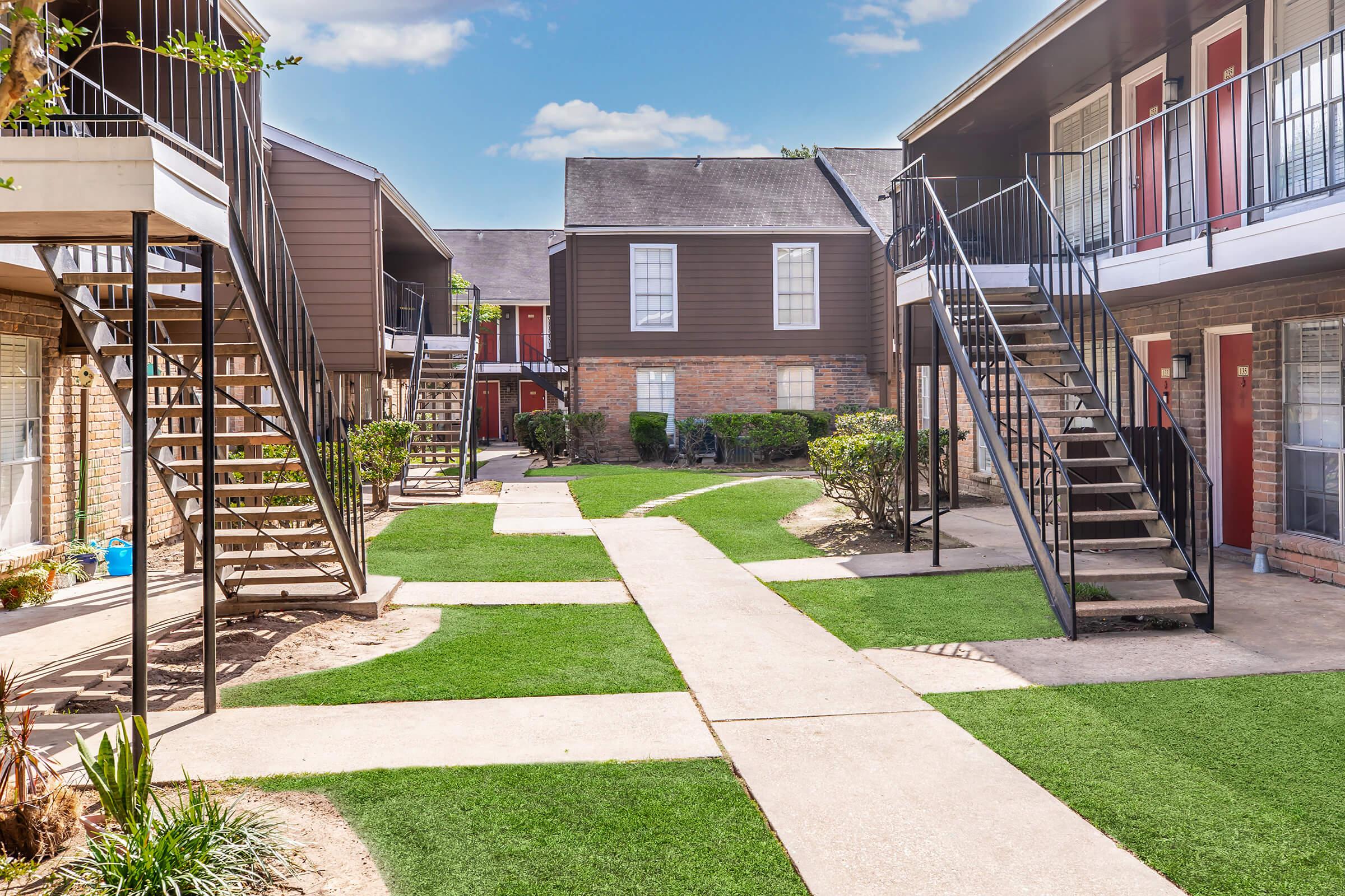 View of a multi-unit apartment complex courtyard, featuring two sets of stairs leading to the upper floors. The area is landscaped with green grass, shrubs, and a gravel path connecting the units. The buildings have a dark exterior with red doors, creating a cozy, inviting atmosphere.