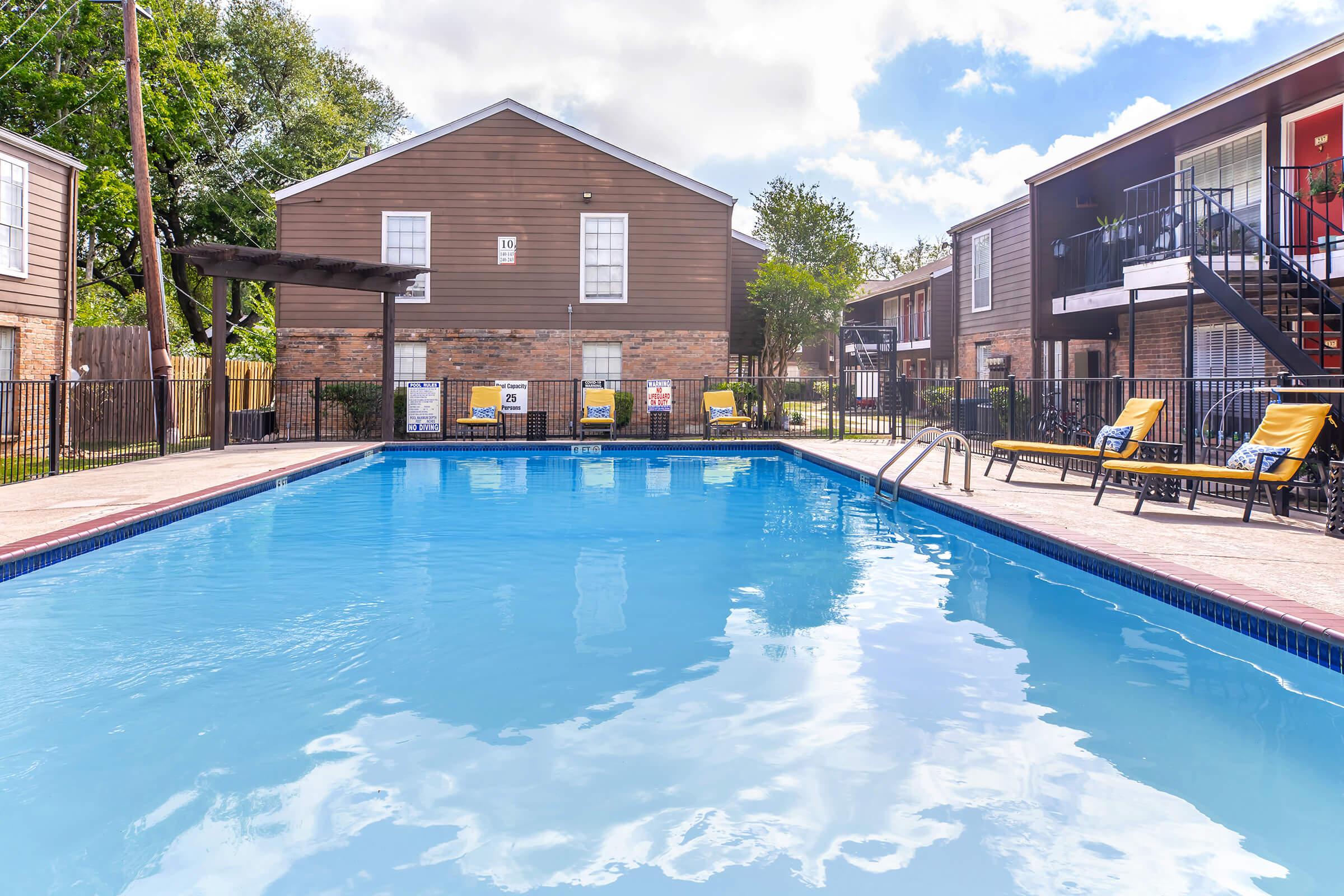 A clear blue swimming pool surrounded by a patio with yellow lounge chairs. In the background, there are two-story residential buildings with brown exteriors, along with trees and blue skies. The scene is inviting and perfect for relaxation.