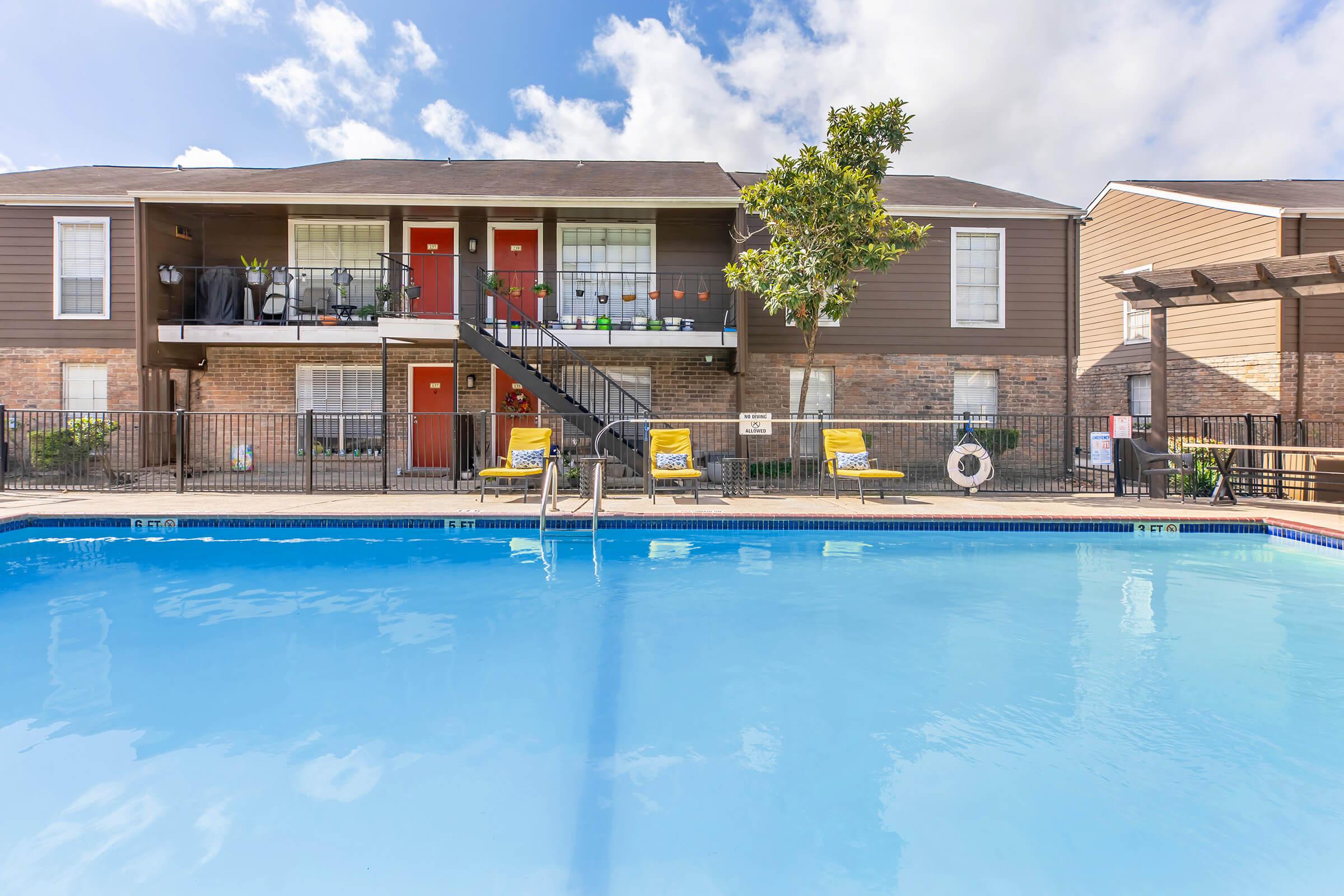 A view of an apartment complex with two-story buildings. A clear blue swimming pool in the foreground, with yellow lounge chairs around it. There are several potted plants and a staircase leading to the upper level of the apartments, set against a backdrop of partly cloudy blue skies.