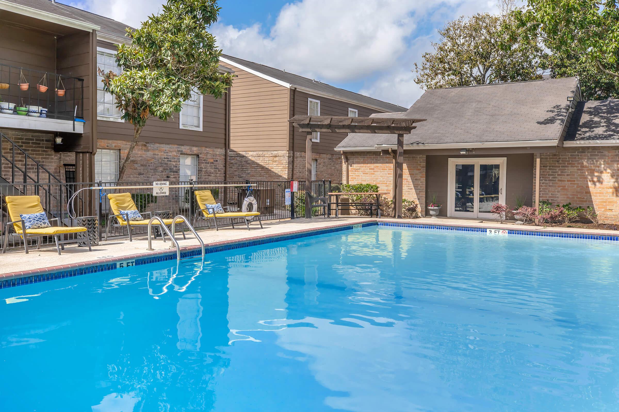 A clear blue swimming pool surrounded by lounge chairs under a sunny sky. In the background, there are two multi-story residential buildings, with one featuring a pergola and decorative plants. The setting is inviting and suitable for relaxation.