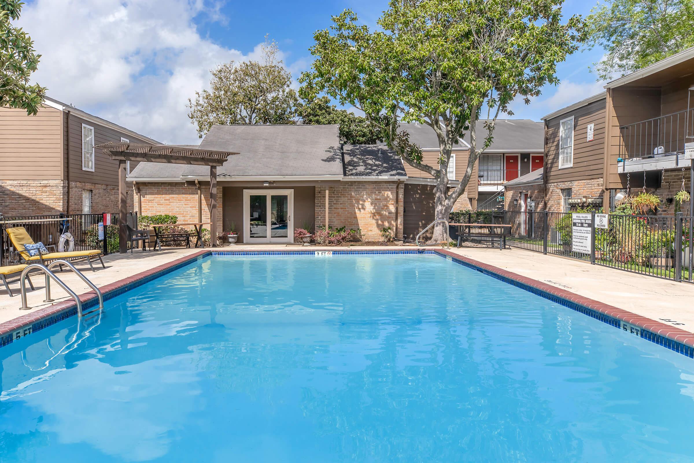 A clear blue swimming pool surrounded by a landscaped area, featuring lounge chairs and trees. In the background, there are residential buildings with a porch area, and a cloudy blue sky overhead. The setting conveys a relaxing outdoor environment.