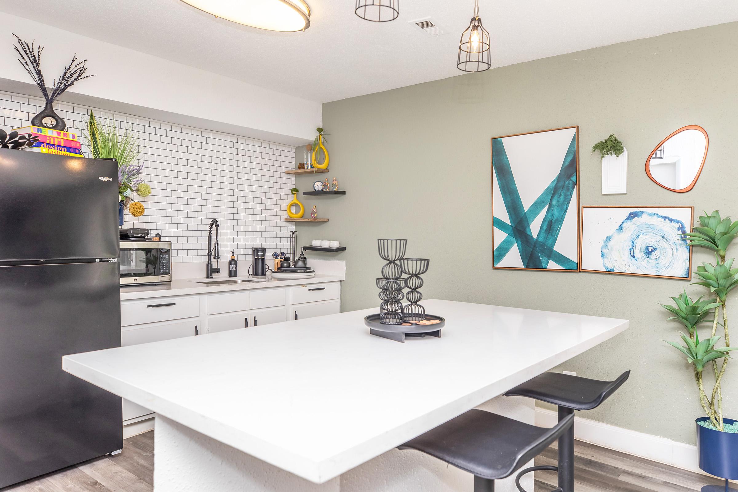 A modern kitchen featuring a white island countertop with black bar stools, a black refrigerator, and a microwave. The walls are painted in pale green and adorned with two framed artworks. There are decorative items on shelves, plants, and a candle holder on the table, creating a stylish and inviting atmosphere.