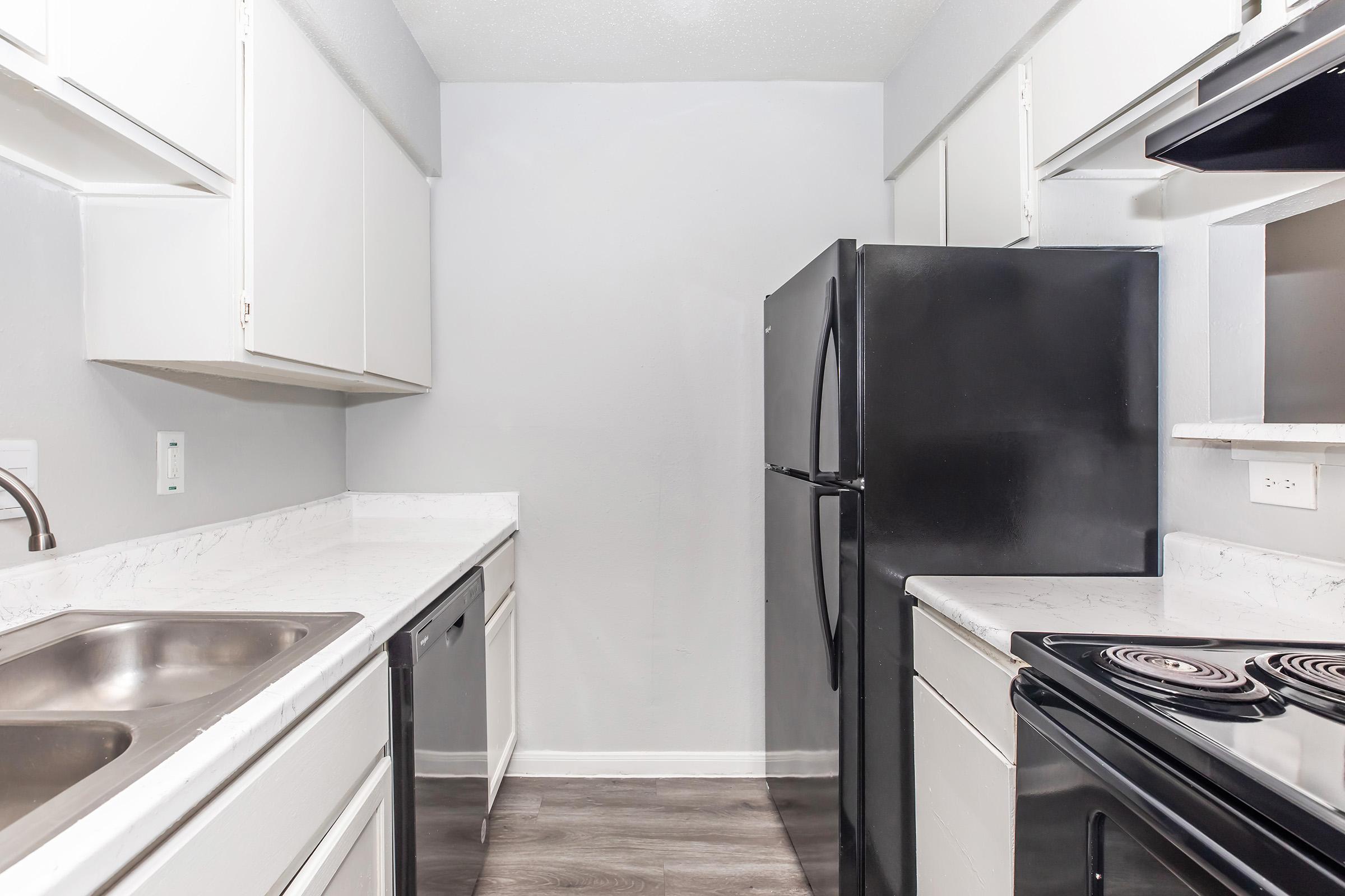 A modern kitchen featuring white cabinets, a black refrigerator, and a black stove. The countertops are light-colored with a marble-like finish. Two sinks are visible, and the kitchen has a clean and organized layout with laminate flooring.