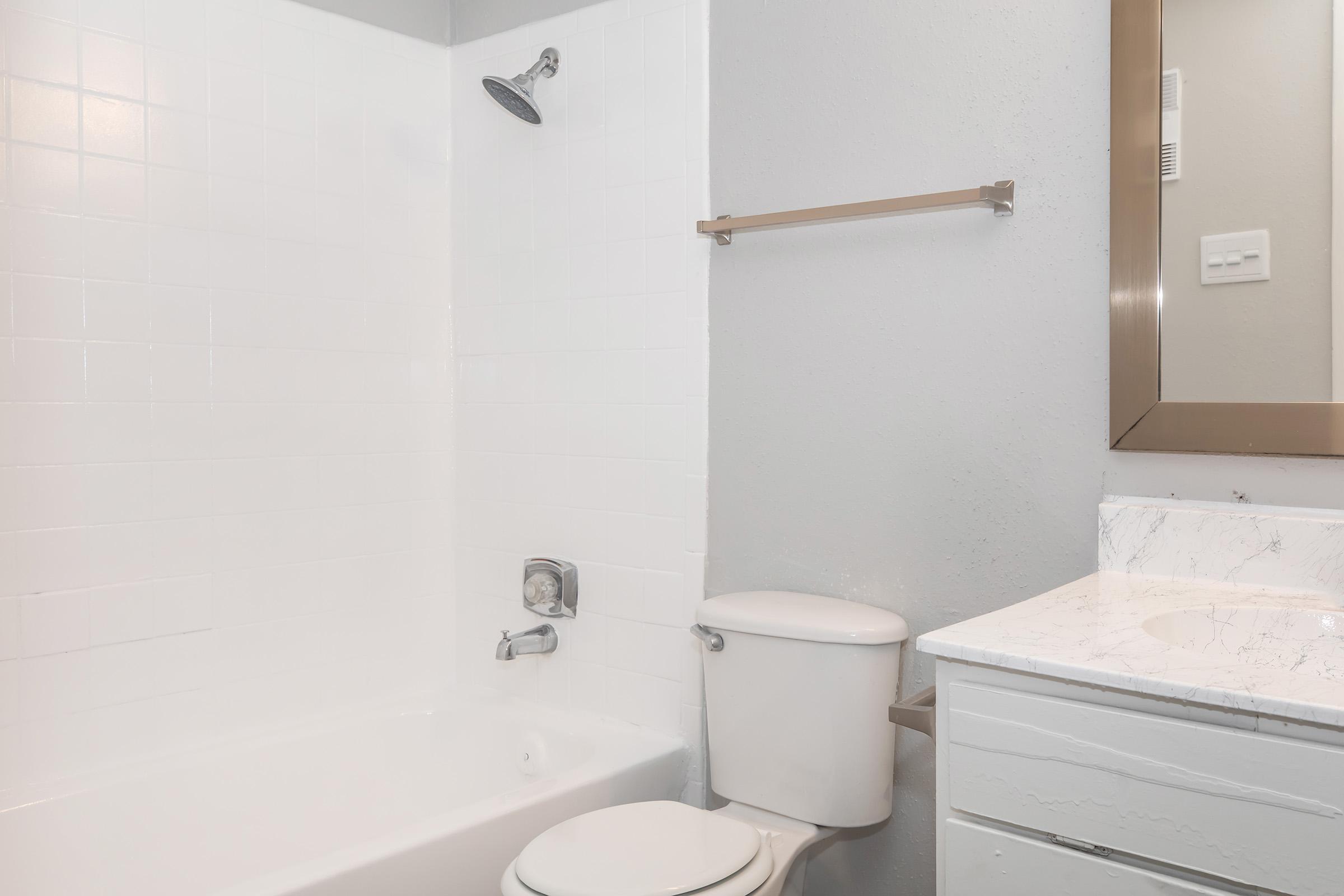 A clean, modern bathroom featuring a white tiled shower with a showerhead, a white bathtub, a toilet, and a sink with a marble countertop. The walls are painted in a light gray color, and there is a mirror above the sink with a small light fixture. A towel rack is mounted on the wall.
