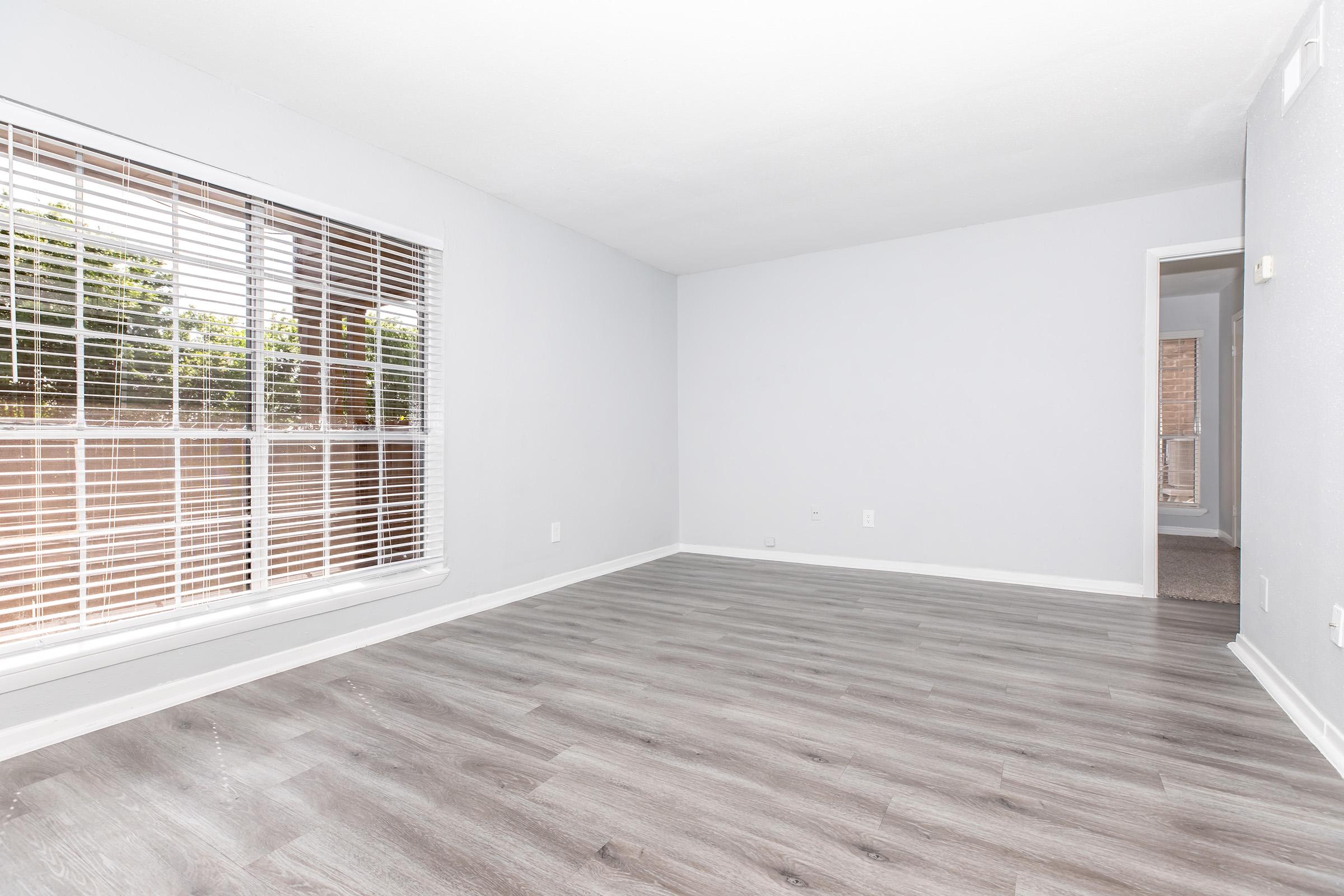 A spacious, light-filled room with gray walls and wood-look flooring. Large windows with white blinds allow natural light to enter, while a doorway leads to another room. The space appears empty, conveying a sense of openness and potential for decoration.