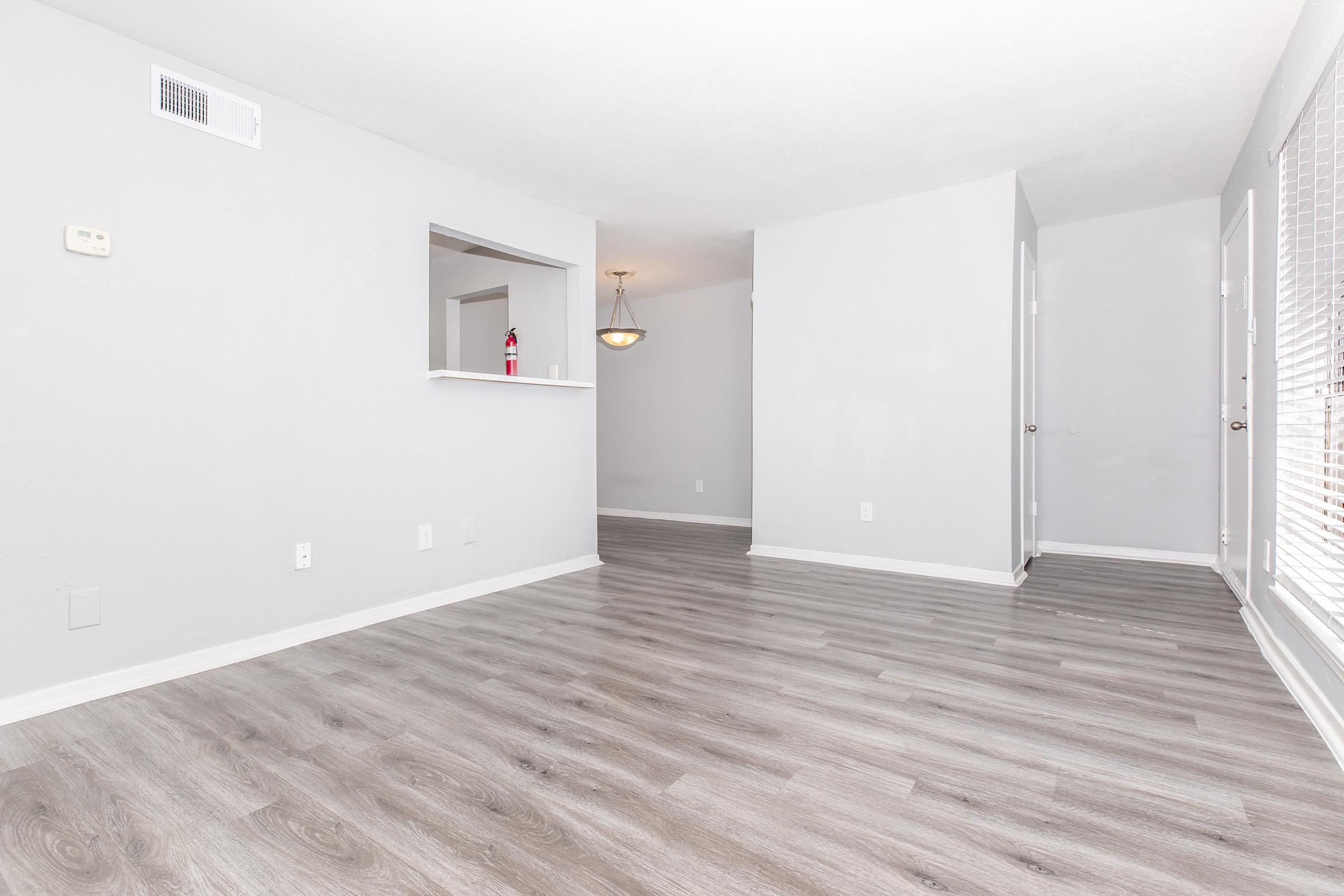 Spacious light-filled living area with gray walls, hardwood-style flooring, and a small serving bar connecting to an adjacent room. A chandelier hangs in the distance, while large windows potentially offer natural light. The overall atmosphere is clean and modern, ideal for a cozy home.