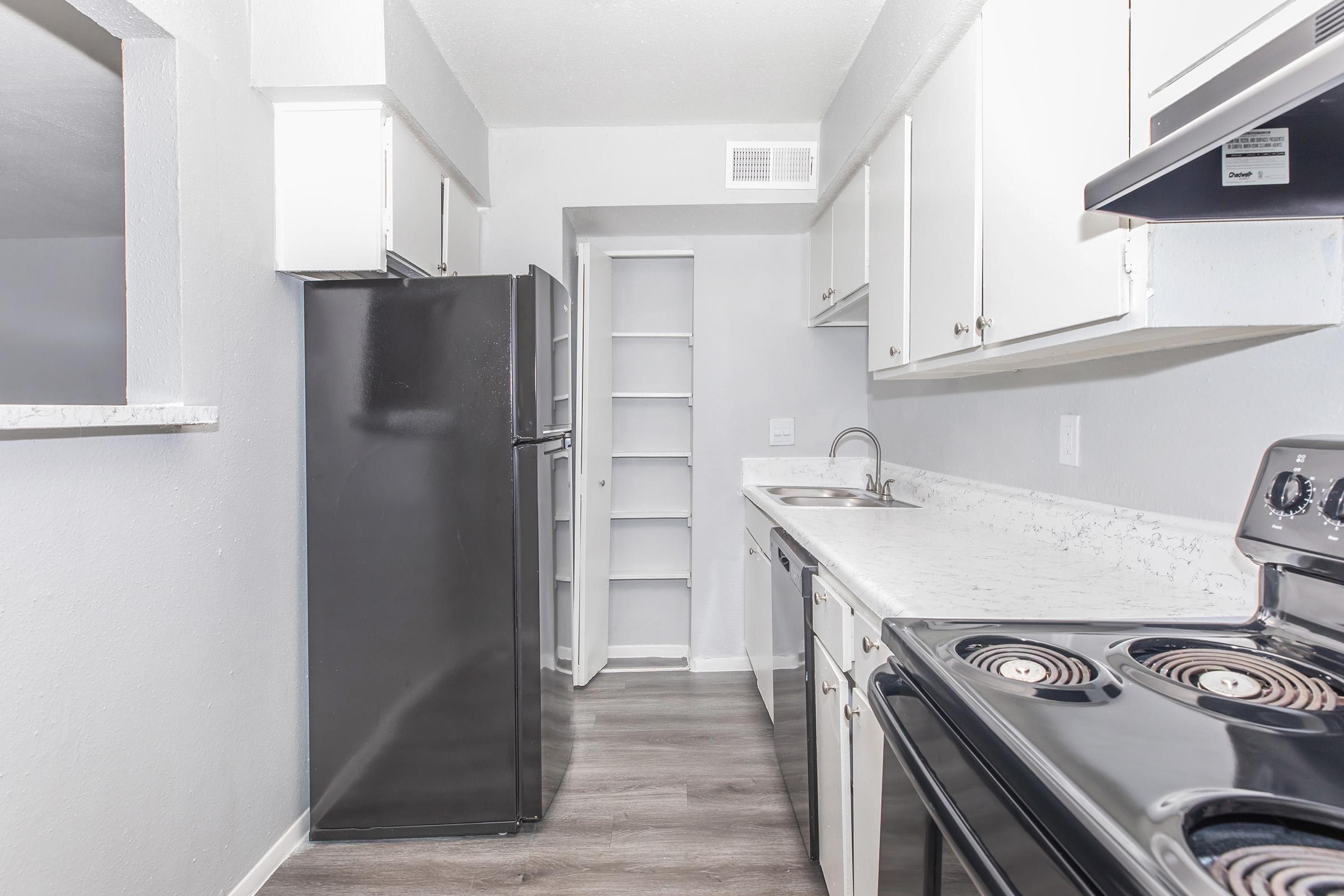 Modern kitchen featuring white cabinets, a black refrigerator, and a stove. The countertop is light-colored stone, with a sink and a small storage area. The walls are painted in a light gray, and the flooring is a wood-like laminate. The kitchen is well-lit and organized.