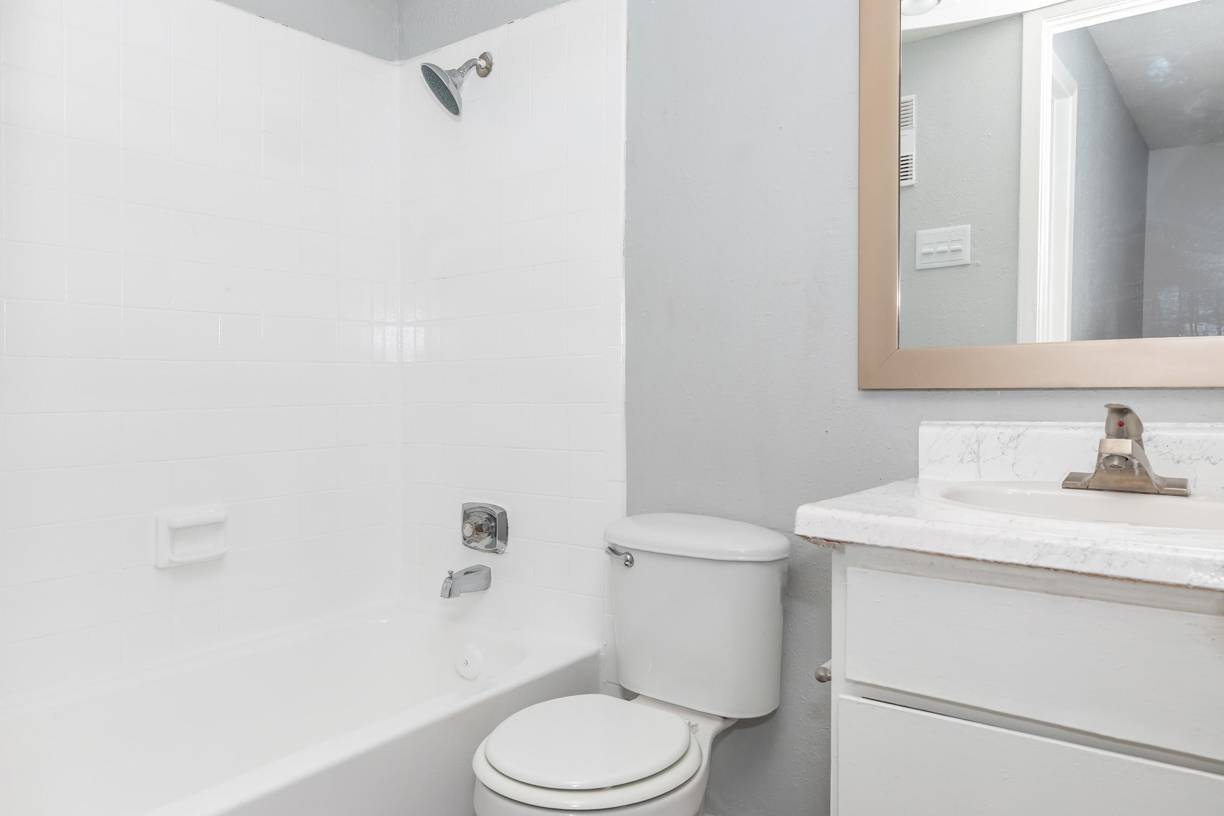A clean and simple bathroom featuring a white bathtub with a showerhead, a toilet, and a sink with a countertop. The walls are painted gray, and there is a mirror above the sink. The overall design is modern and minimalistic, offering a fresh and tidy appearance.