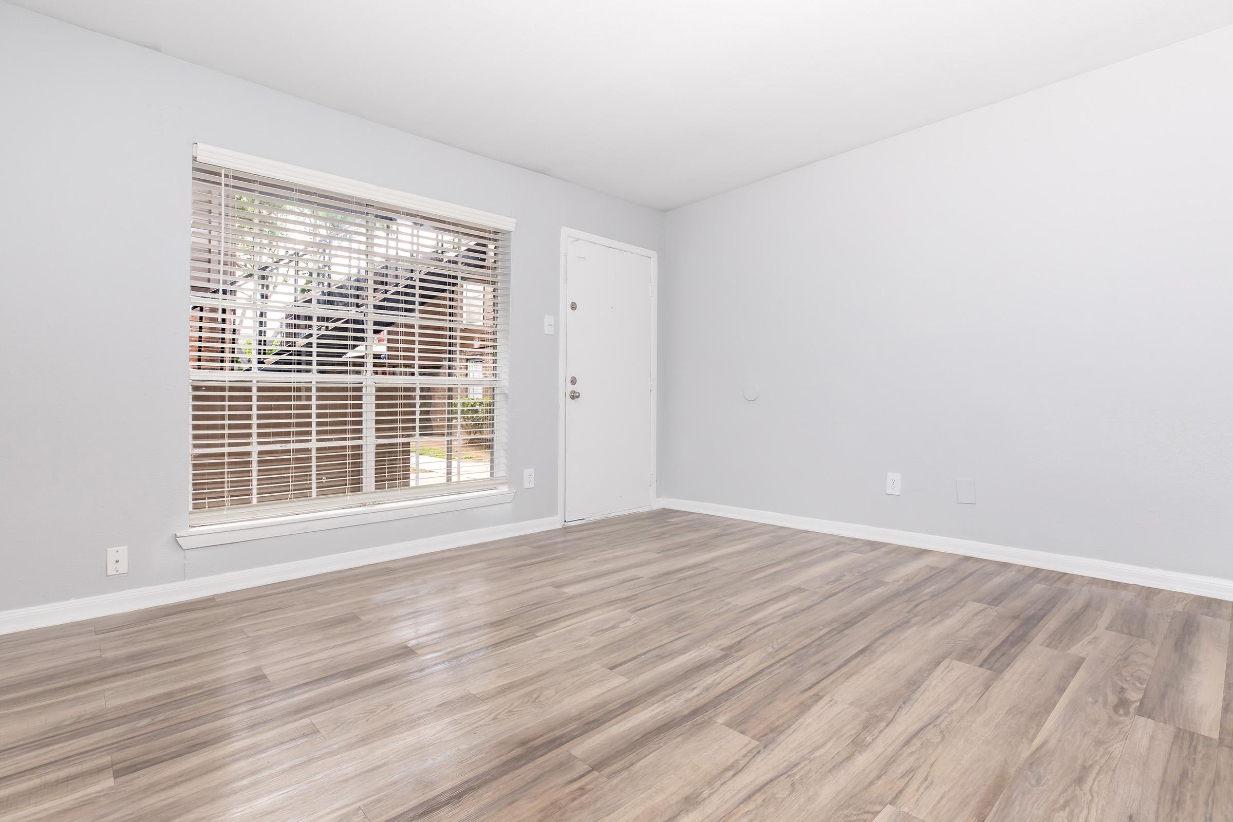 Empty room with light gray walls and a wood-style floor. A window with blinds lets in natural light, providing a view of an outdoor staircase. The door is white and there are no furniture or decorations, creating a minimalist and spacious appearance.