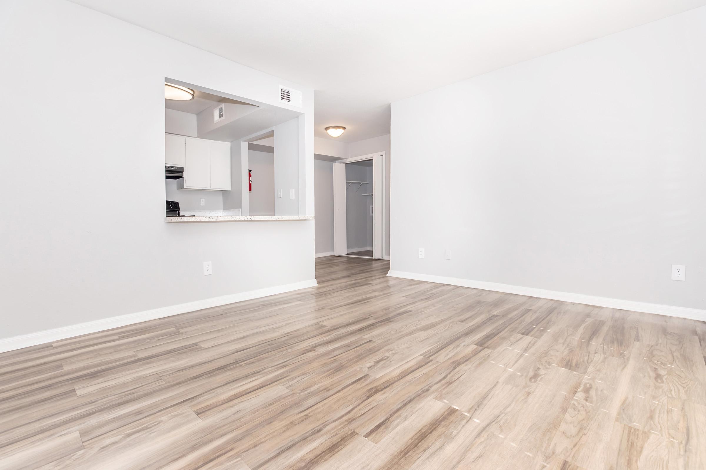 A spacious, empty room with light gray walls and warm wooden flooring. There is a kitchen area visible in the background with white cabinetry. A doorway leads to another room, while ample natural light brightens the space. The overall atmosphere is clean and modern, ideal for furniture arrangement.
