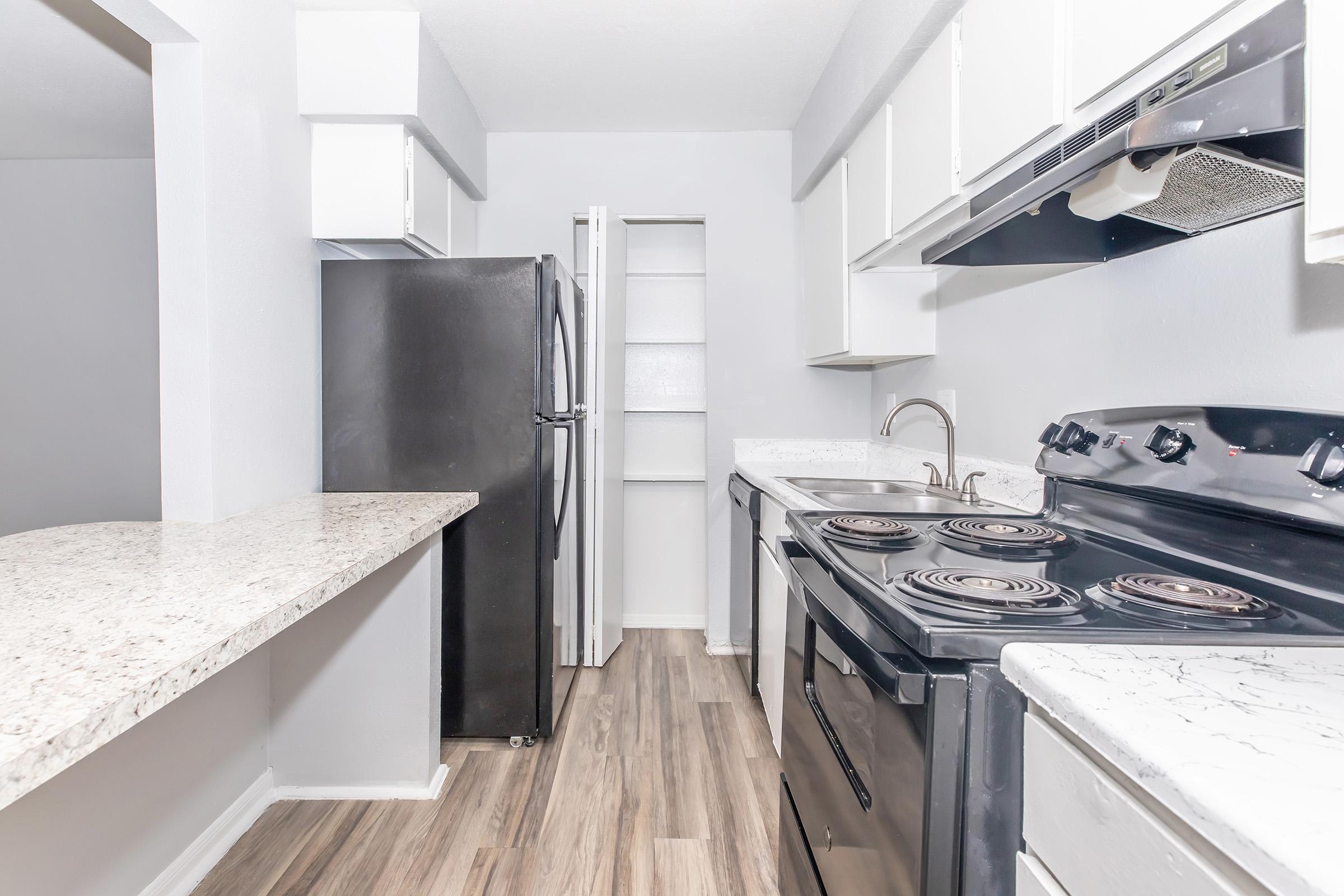 A modern kitchen featuring black appliances, including a refrigerator and stove, with white cabinets and a granite countertop. The flooring is light wood, and there is an open pantry space visible in the background, creating a spacious and functional layout.