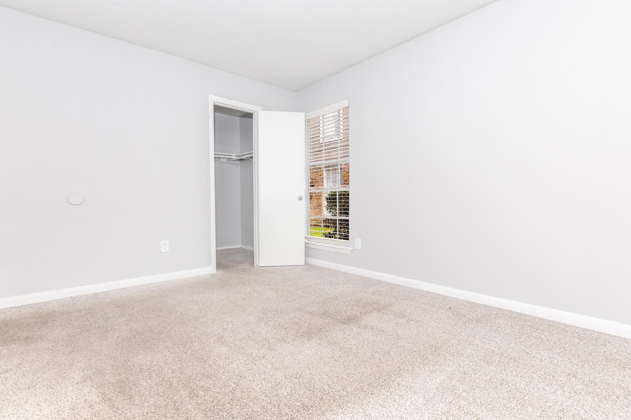 A light-colored empty bedroom with beige carpet, featuring a partially open door leading to a closet and a window with natural light coming in. The walls are painted in a soft gray, and the overall space appears clean and inviting.