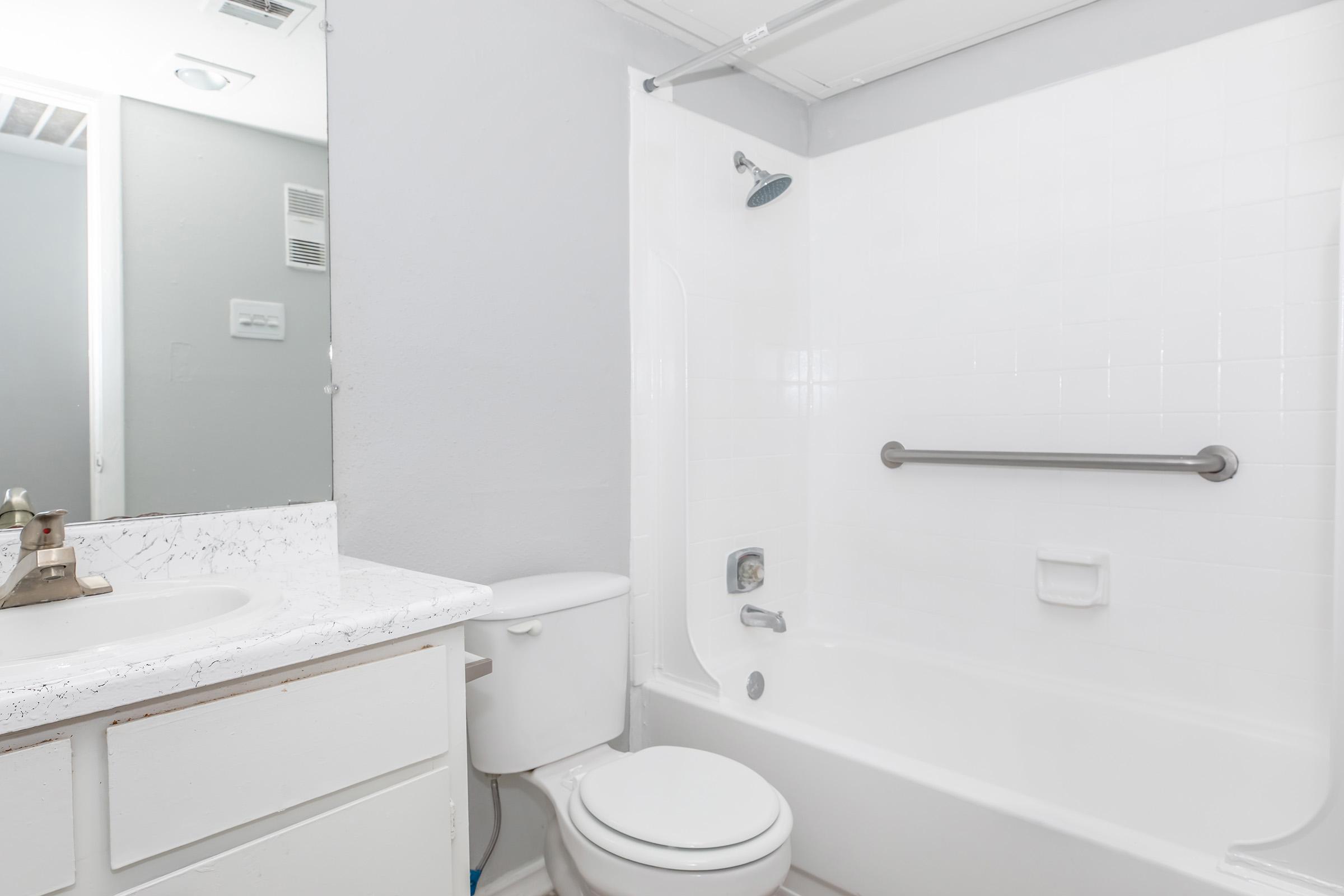 A clean, modern bathroom featuring a white bathtub with a showerhead, a toilet, and a vanity with a sink. The walls are painted light gray, and there is a mirror reflecting the space. The bathroom has minimalistic decor, creating a simple and tidy appearance.