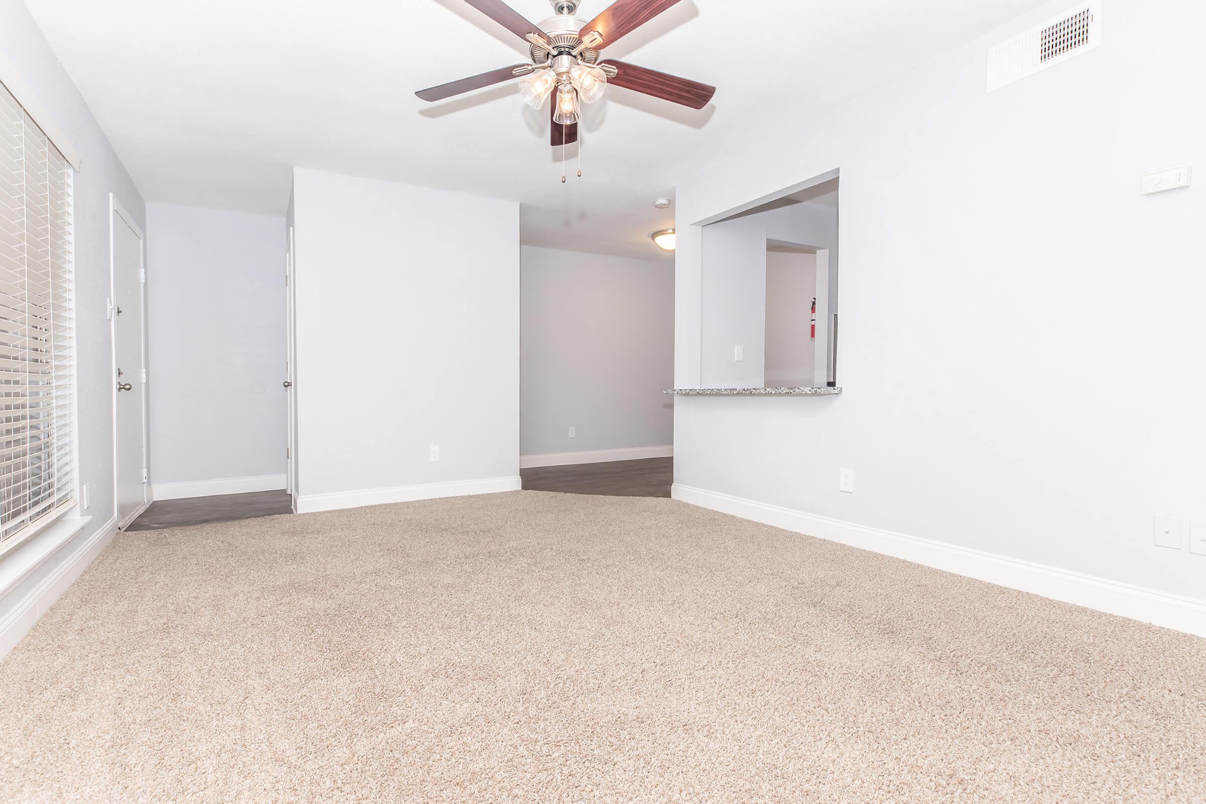 Bright, empty living room featuring light gray walls, a ceiling fan, and plush beige carpeting. A large window with blinds allows natural light to enter, while a doorway leads to another room. The space is clean and ready for furnishings, creating a welcoming atmosphere.
