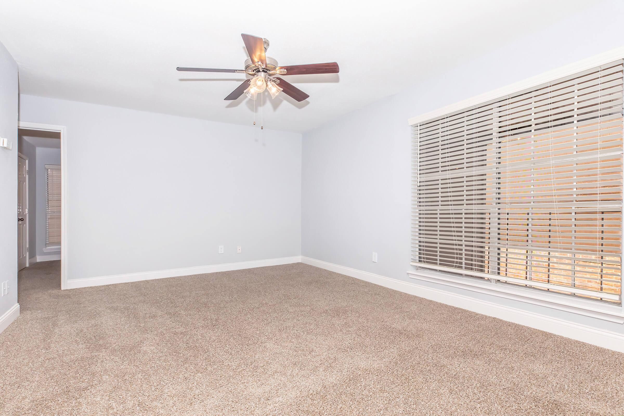A spacious, empty room with light gray walls and a ceiling fan featuring four lights. The floor is covered in beige carpet, and large windows with wooden blinds allow natural light. The room has a cozy and clean appearance, suitable for various uses, such as a living or bedroom space.