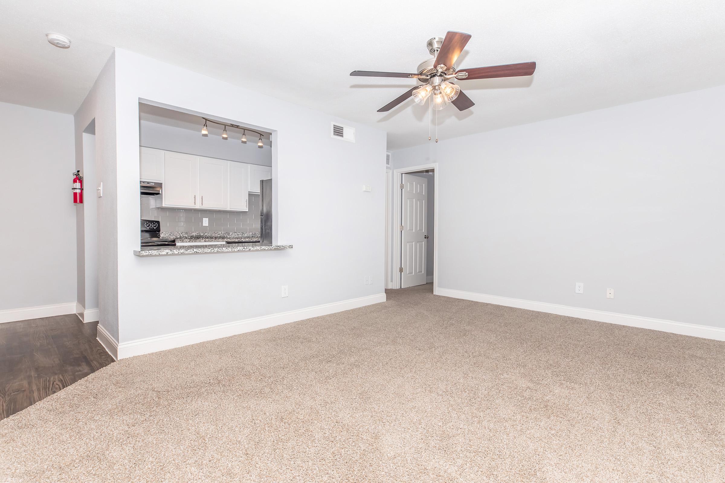 Spacious living area with light-colored walls and plush carpet. A ceiling fan is installed, and a breakfast bar connects to a modern kitchen with pendant lighting. A doorway leads to a closed room, while a fire extinguisher is mounted on the wall, indicating safety features in the space.