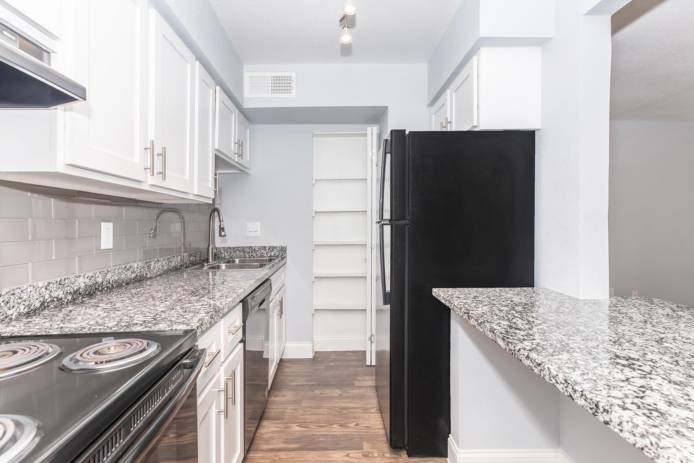 Modern kitchen featuring white cabinets, a black refrigerator, a stainless steel sink, and a granite countertop. The space includes a stove with an oven and an organized pantry shelf visible in the background. The walls are painted light blue, creating a bright and inviting atmosphere.