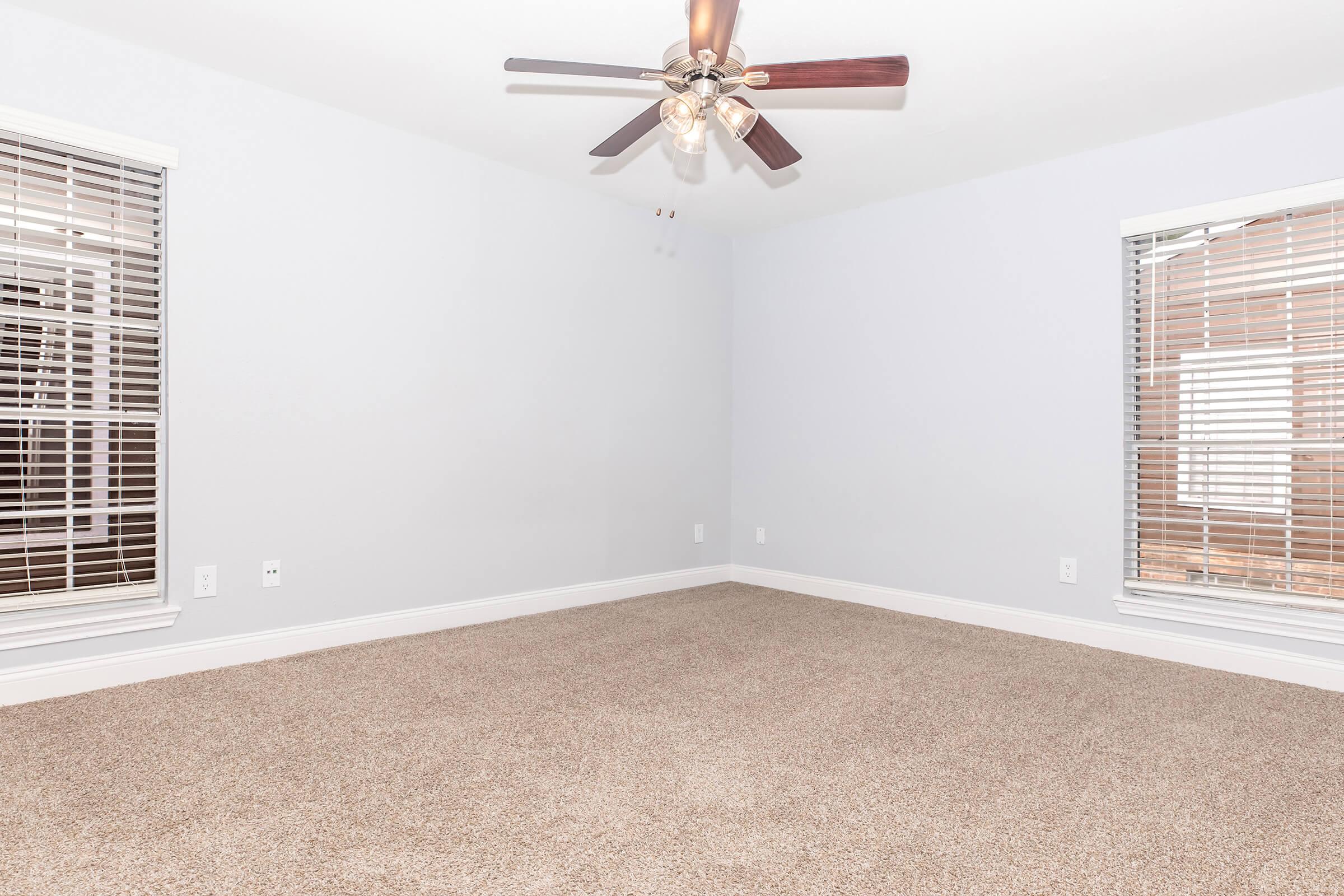 A spacious, empty room with light gray walls and brown carpet. It features a ceiling fan with wooden blades and two windows with blinds, allowing natural light to enter. The room has a clean, minimalist appearance, suitable for various decorating styles.