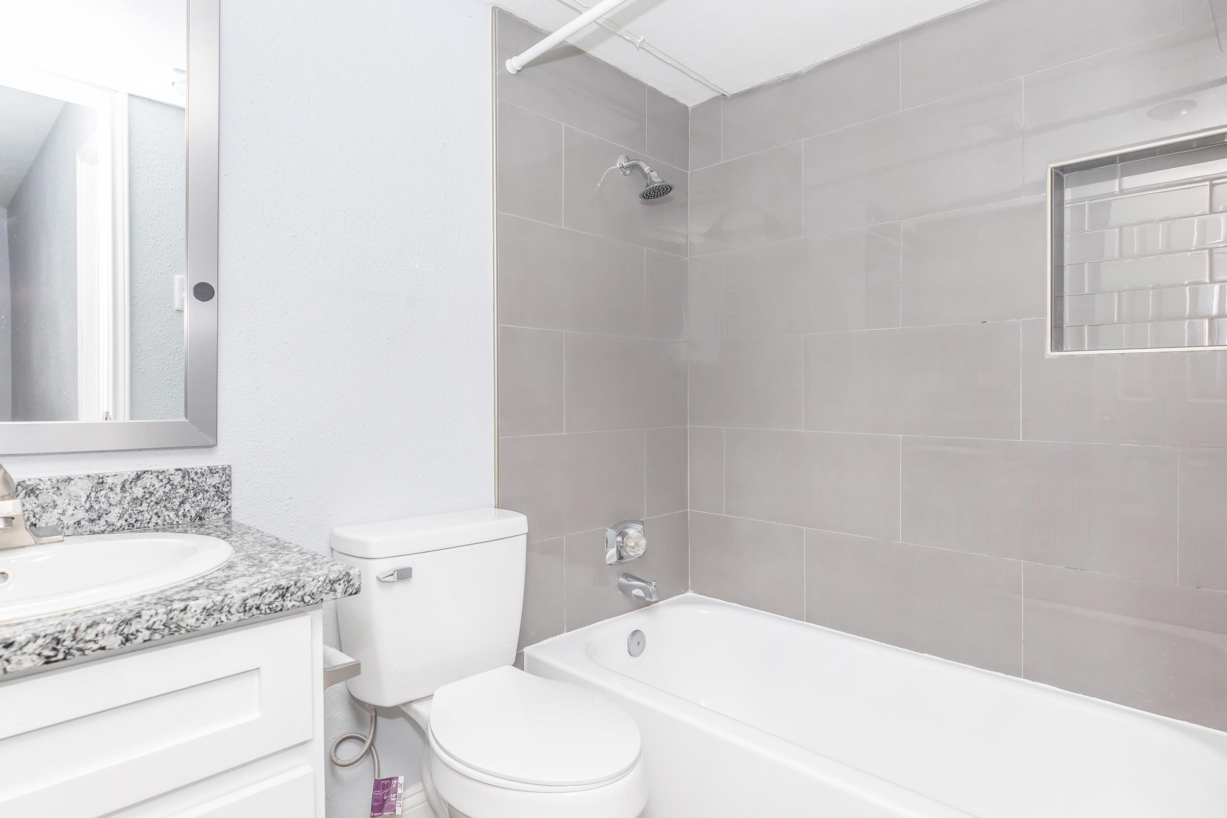 A modern bathroom featuring a white bathtub, toilet, and a granite countertop sink. The walls are covered in light gray tiles, and there's a shower with a glass door. A mirror is visible on the wall, reflecting the space. The overall design is clean and minimalistic.