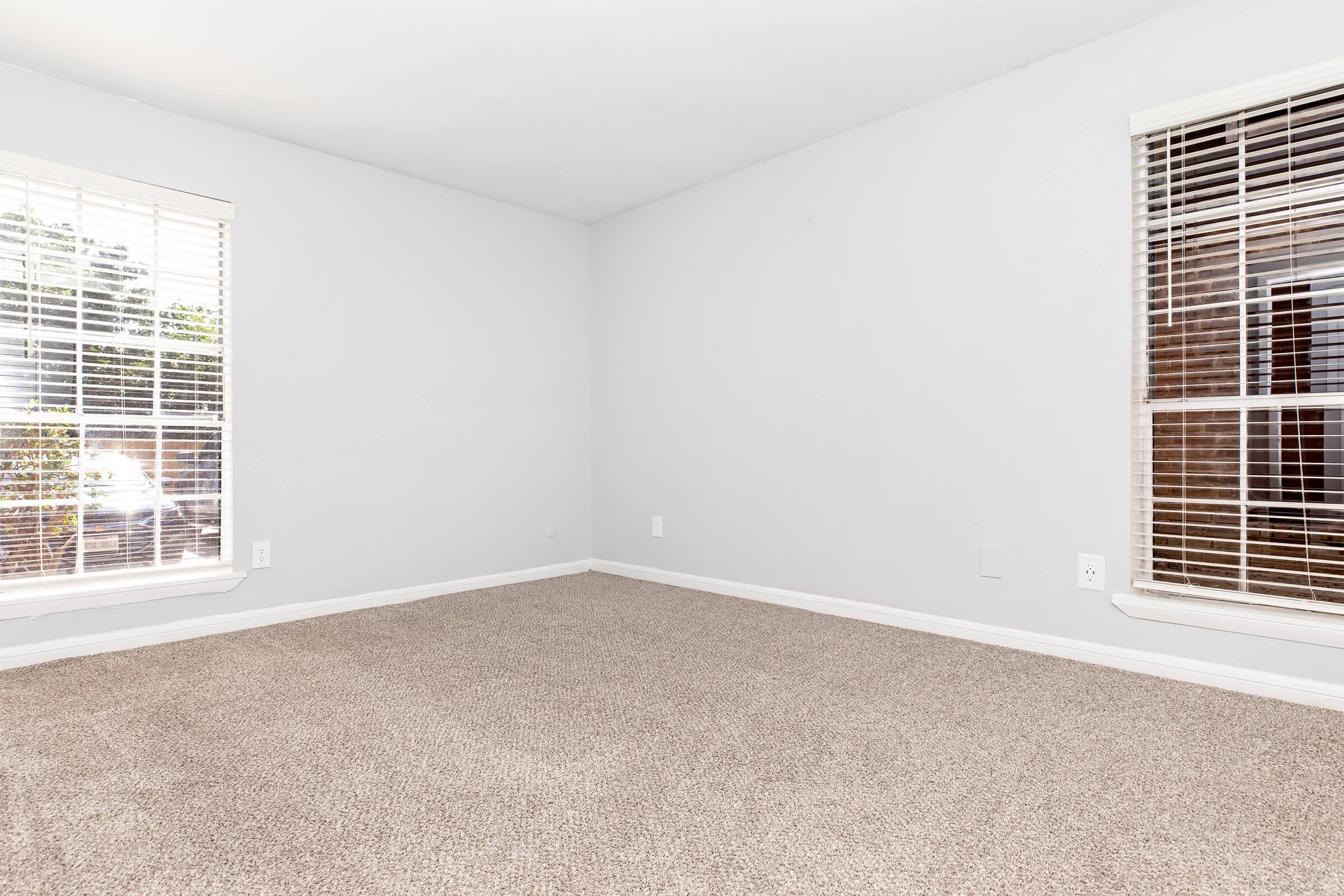A spacious, empty room with light gray walls and brown carpet. Two windows with white blinds allow natural light to enter, creating a bright atmosphere. The room has a clean and minimalistic design, making it suitable for various purposes like a living area or office space.