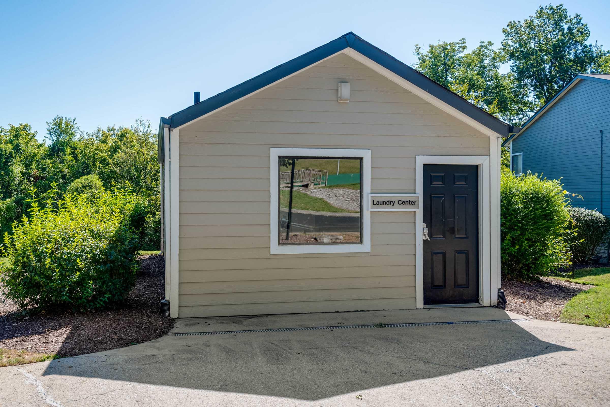 Exterior view of a small laundry center building with a dark front door and a window. The building is light-colored with a sign labeled "Laundry Center." Surrounding the building are well-kept shrubs and greenery, set against a clear blue sky. The area appears tidy and inviting.