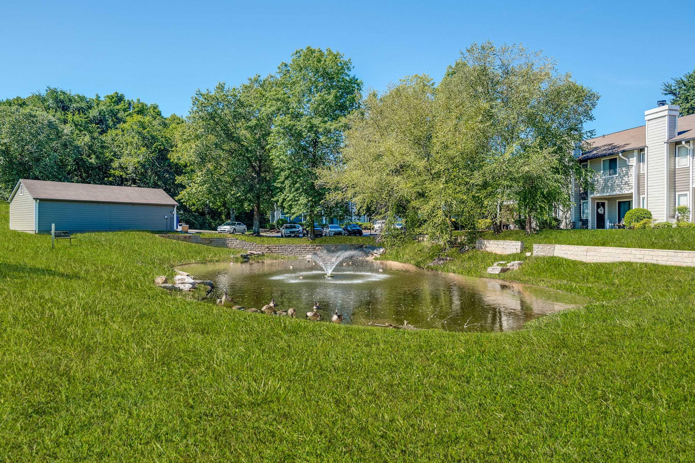 A serene residential landscape featuring a small pond with a fountain surrounded by lush green grass. Trees line the background, and there are townhome units nearby, creating a peaceful, inviting atmosphere. The clear blue sky enhances the tranquil setting.