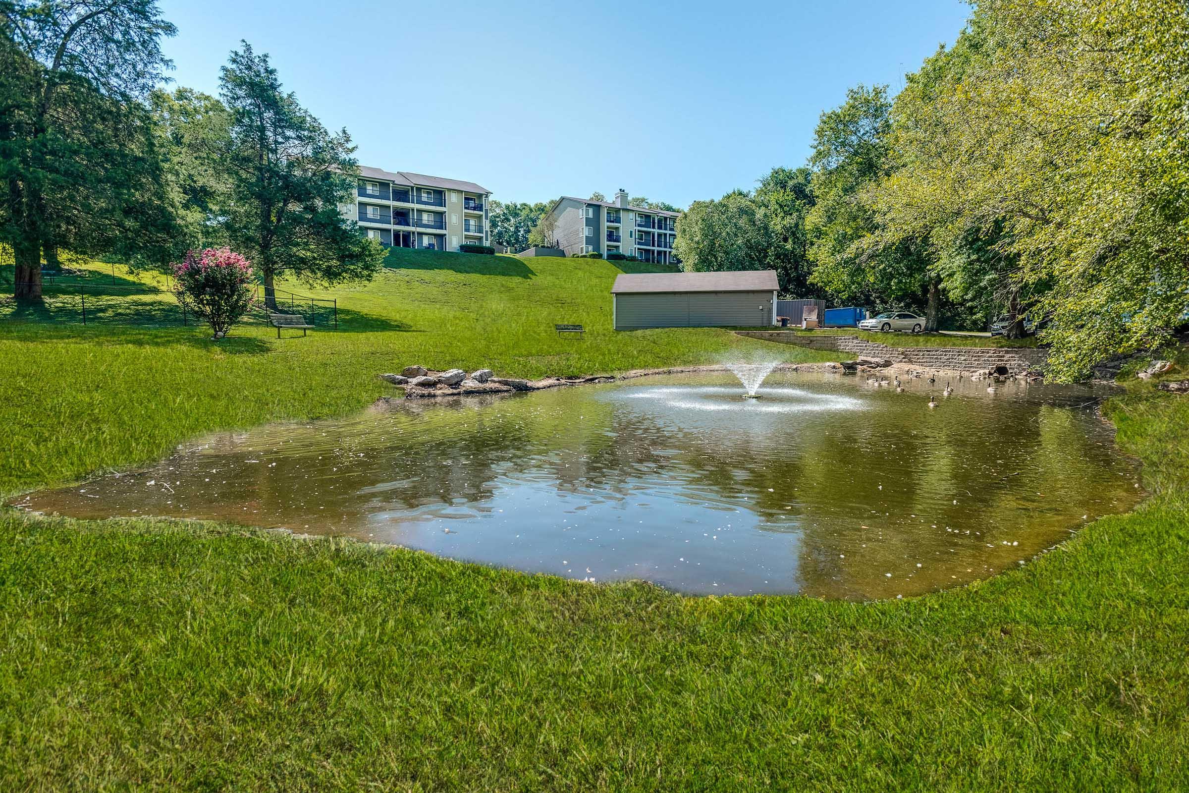 A serene landscape featuring a pond with a fountain, surrounded by lush green grass. In the background, there are several multi-story residential buildings nestled among trees, creating a peaceful, natural setting. The sky is clear and blue, enhancing the tranquil atmosphere of the scene.