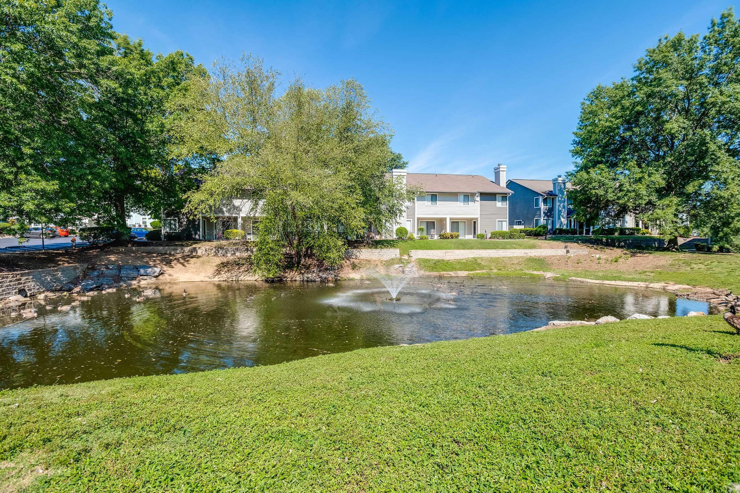 A serene landscape featuring a small pond with a fountain, surrounded by lush green grass and trees. In the background, there are several residential buildings, providing a peaceful and attractive living environment. The sky is clear and blue, enhancing the natural beauty of the scene.