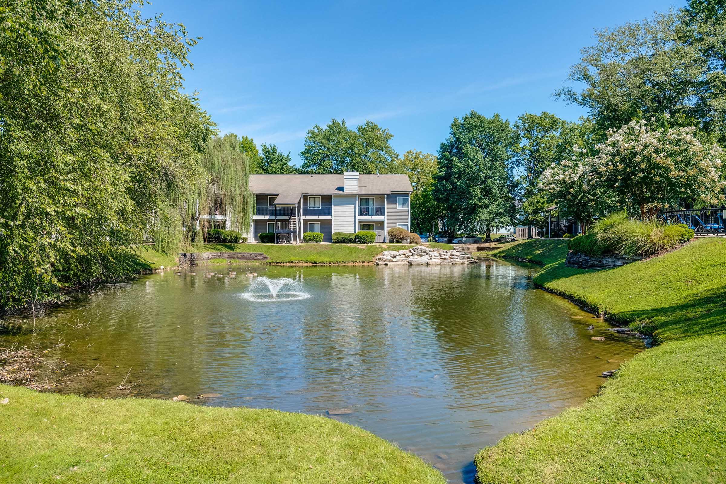 A scenic view of a serene pond surrounded by lush greenery and trees, with a fountain gently spraying water. In the background, there are apartment buildings with balconies, situated near the water's edge and framed by vibrant landscaping under a bright blue sky.