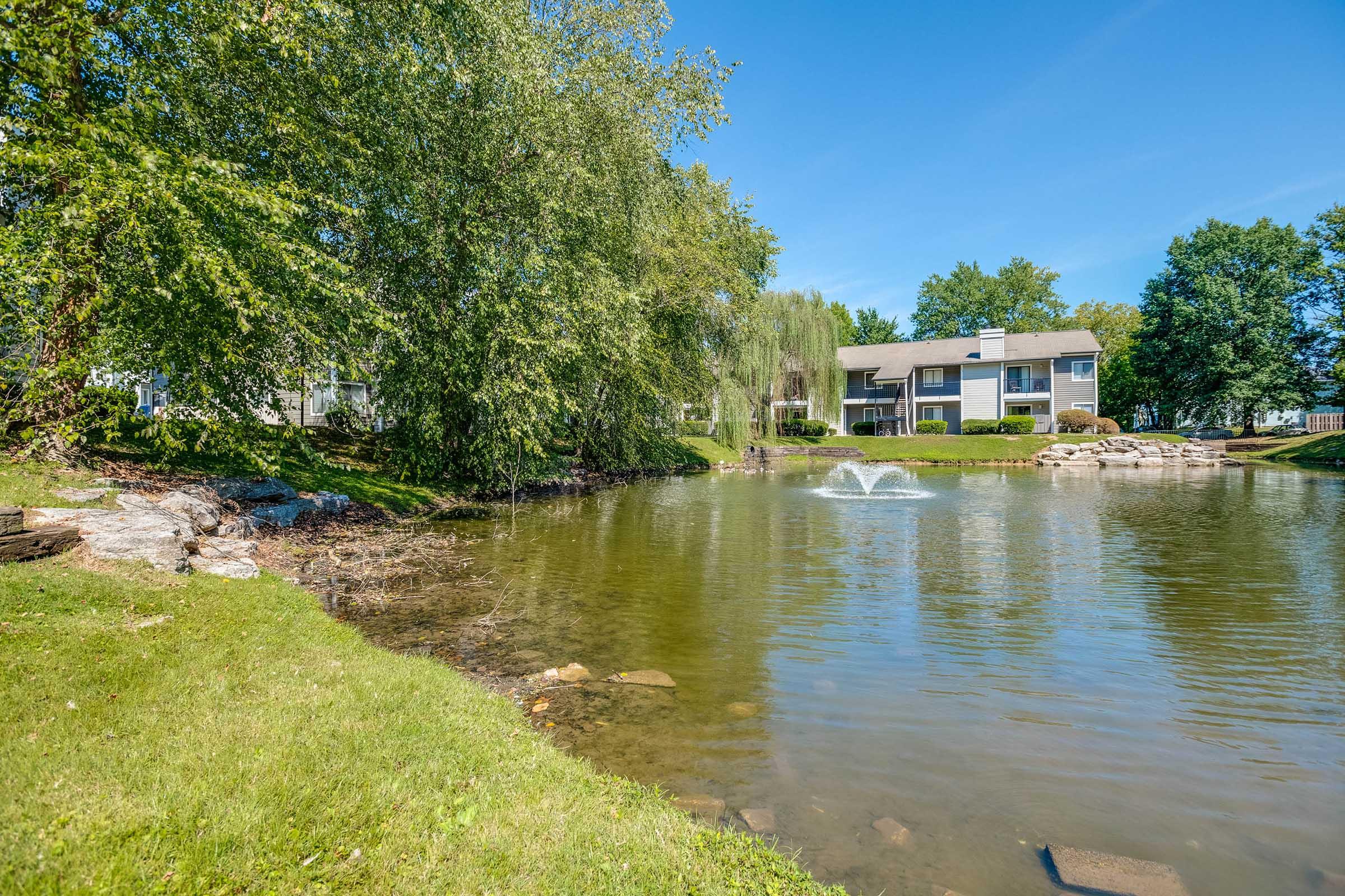 A serene pond surrounded by lush greenery and trees, with a fountain gently spraying water in the center. In the background, a two-story residential building is visible along the shore, framed by well-maintained landscaping under a clear blue sky.