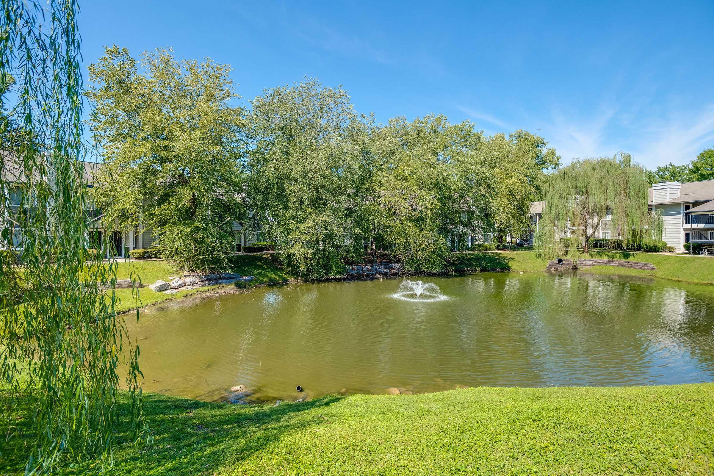 A serene landscape featuring a pond with a fountain, surrounded by lush greenery and trees. In the background, there are residential buildings, partially obscured by vegetation, under a clear blue sky. The scene conveys a tranquil and peaceful atmosphere.
