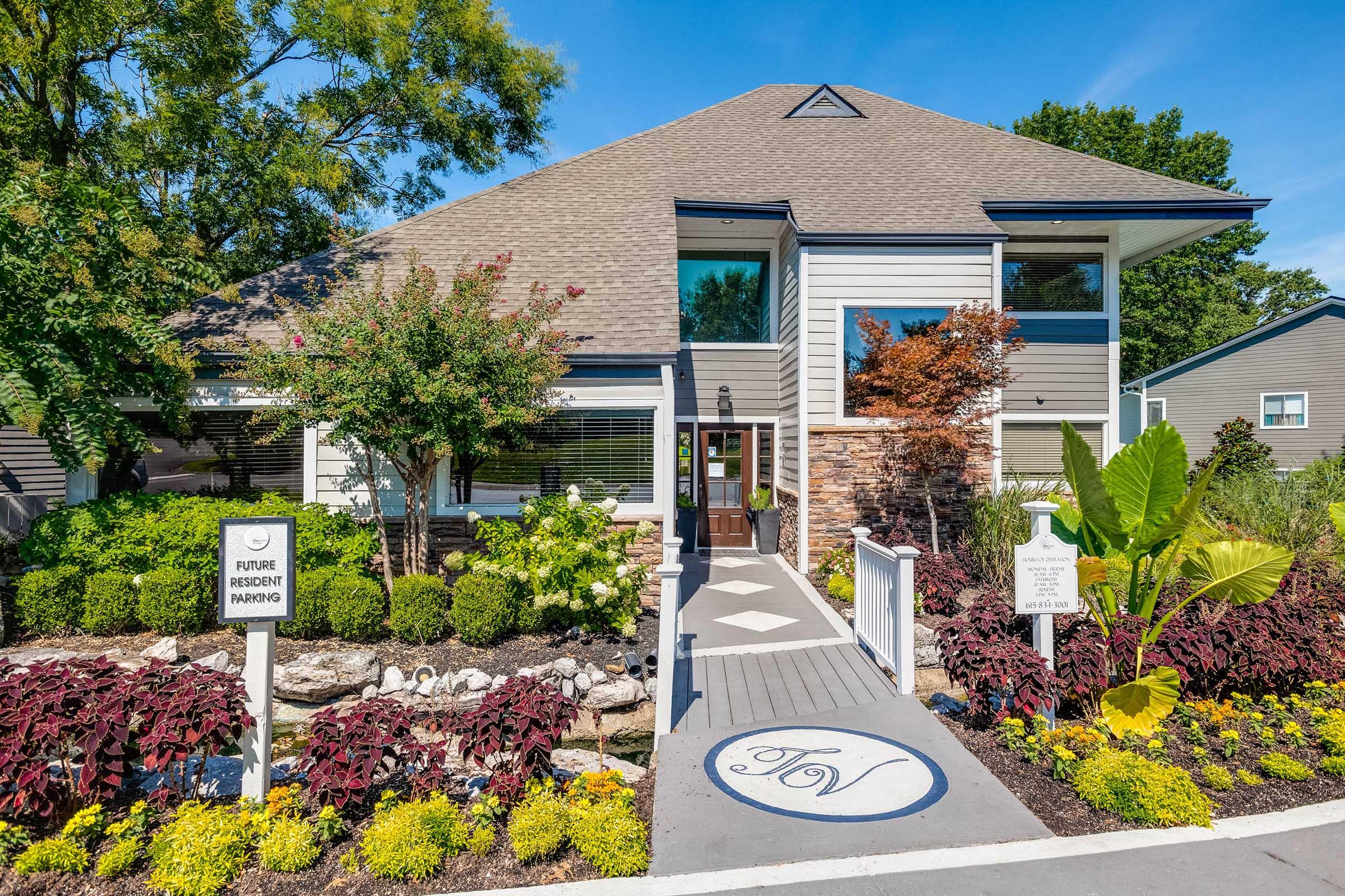 A modern, two-story building with a grey and white exterior features a prominent entrance, landscaped gardens with colorful flowers and shrubs. There are signs indicating future resident parking. The sky is clear and blue, adding to the welcoming atmosphere of the property.