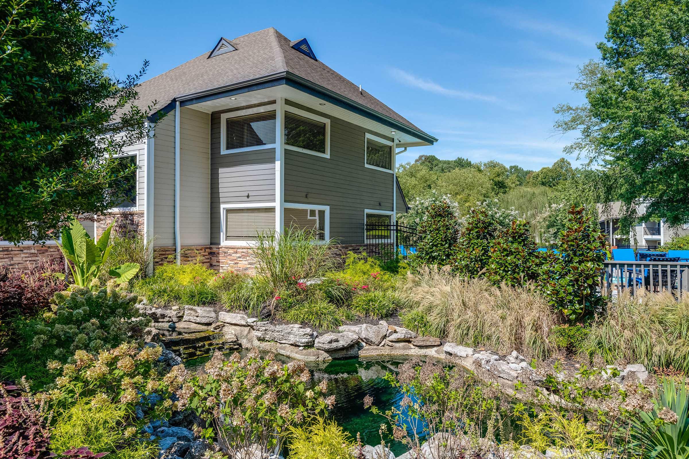 A modern two-story house with a sloped roof, featuring large windows. The surrounding landscape includes lush greenery, decorative shrubs, and a serene pond with rocks. The clear blue sky complements the vibrant foliage, creating a peaceful outdoor setting.