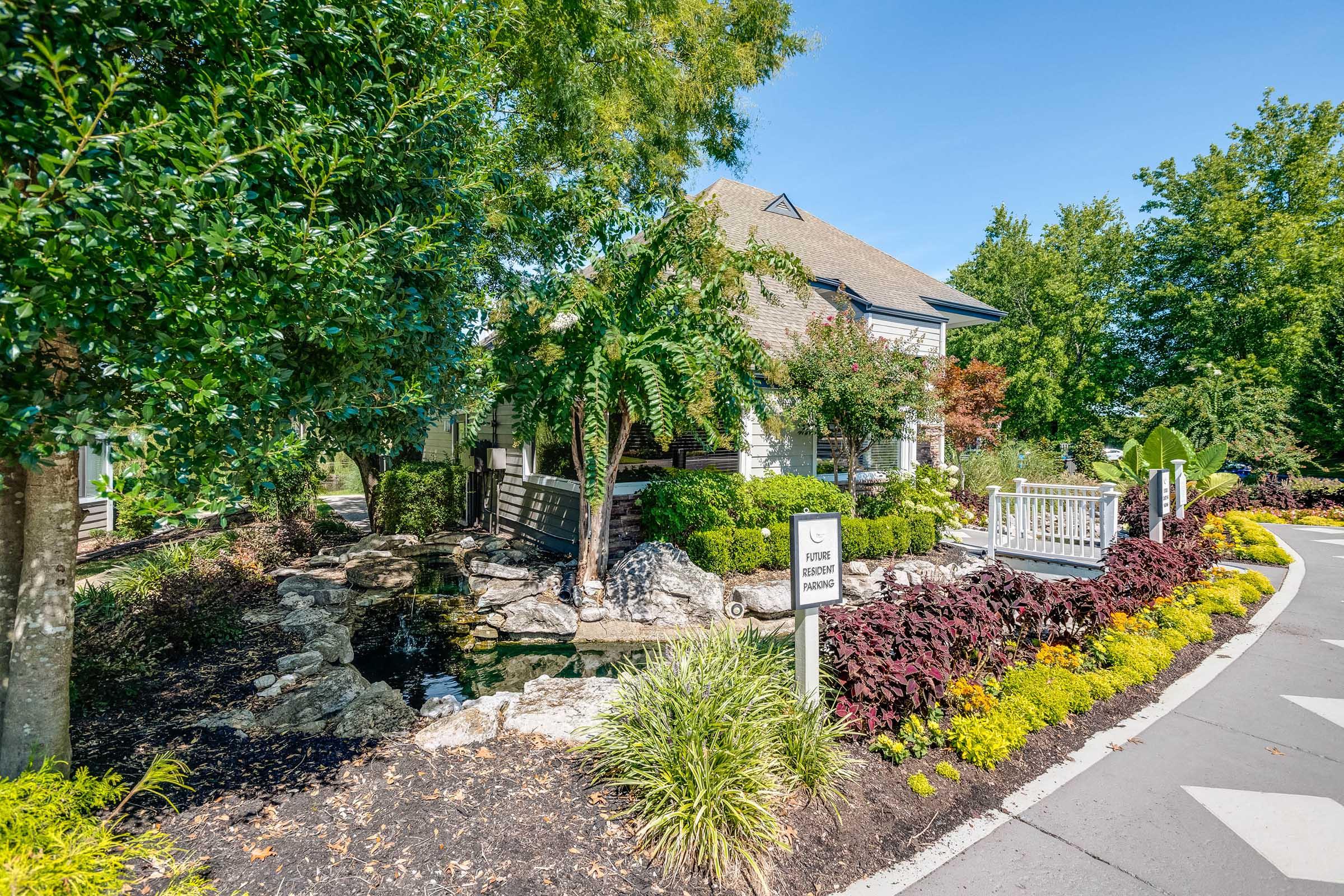 A well-manicured landscape featuring a stone pathway, lush greenery, and colorful flower beds. In the background, there is a charming building with a porch, surrounded by trees. A small pond is visible among the plants, enhancing the serene outdoor atmosphere.