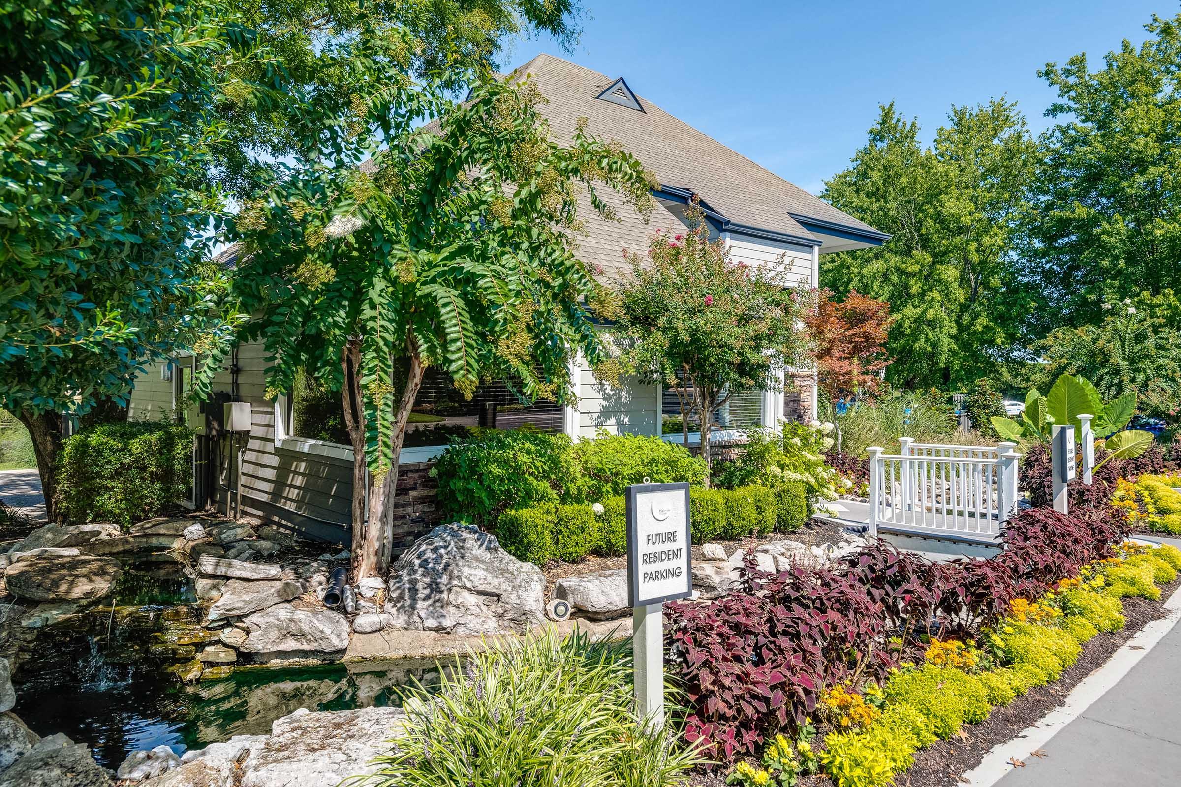 A picturesque house surrounded by lush greenery and colorful flowers, featuring a small pond with rocks. A sign in front indicates “Future Resident Parking.” The scene is bright and sunny, showcasing a well-maintained landscape with trees and shrubs enhancing the environment.