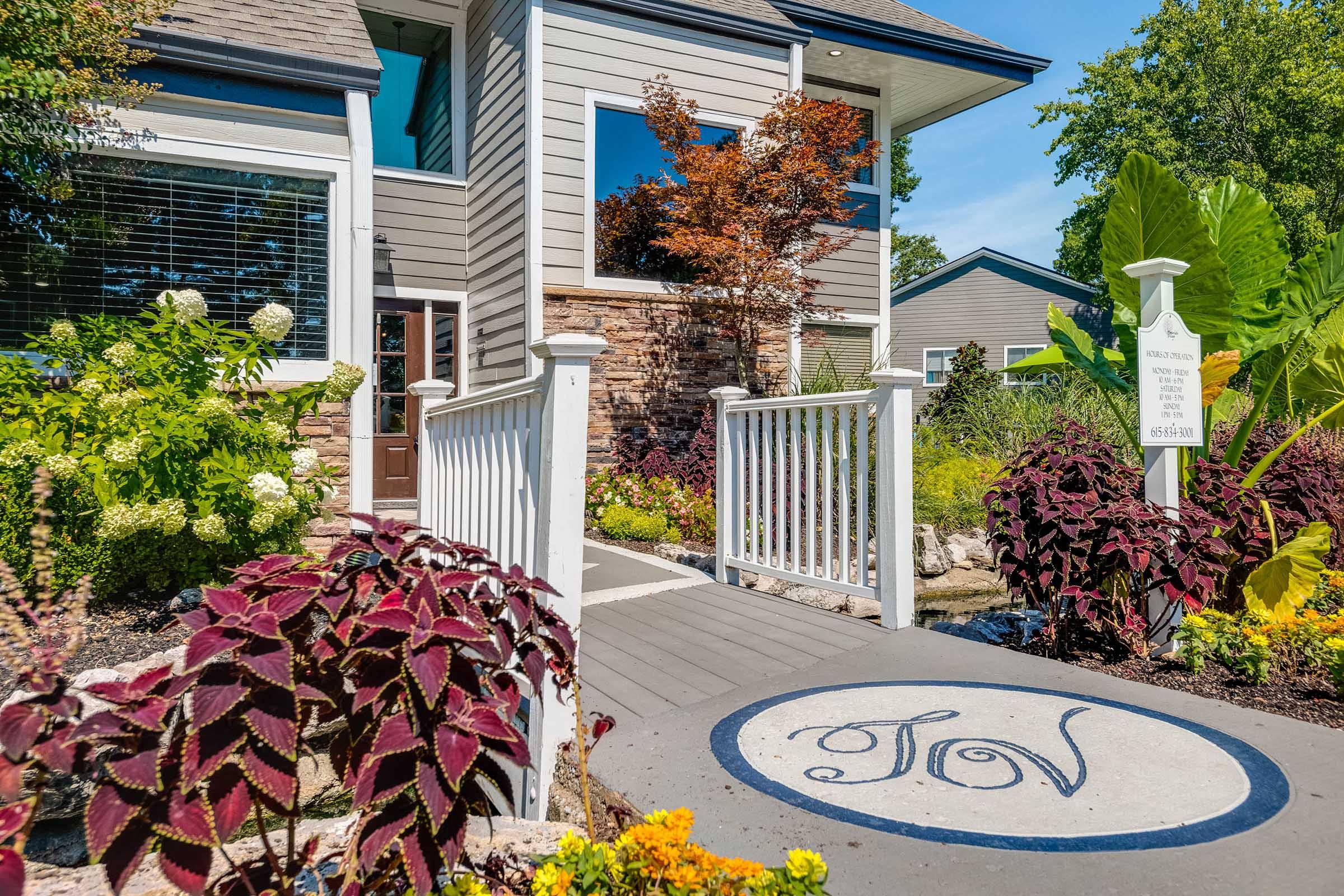 A well-maintained garden with colorful plants and flowers leads to a modern home. The pathway features a circular emblem with "JV" in the center, bordered by lush greenery. The house has large windows and a welcoming front porch, creating an inviting atmosphere in a residential neighborhood.