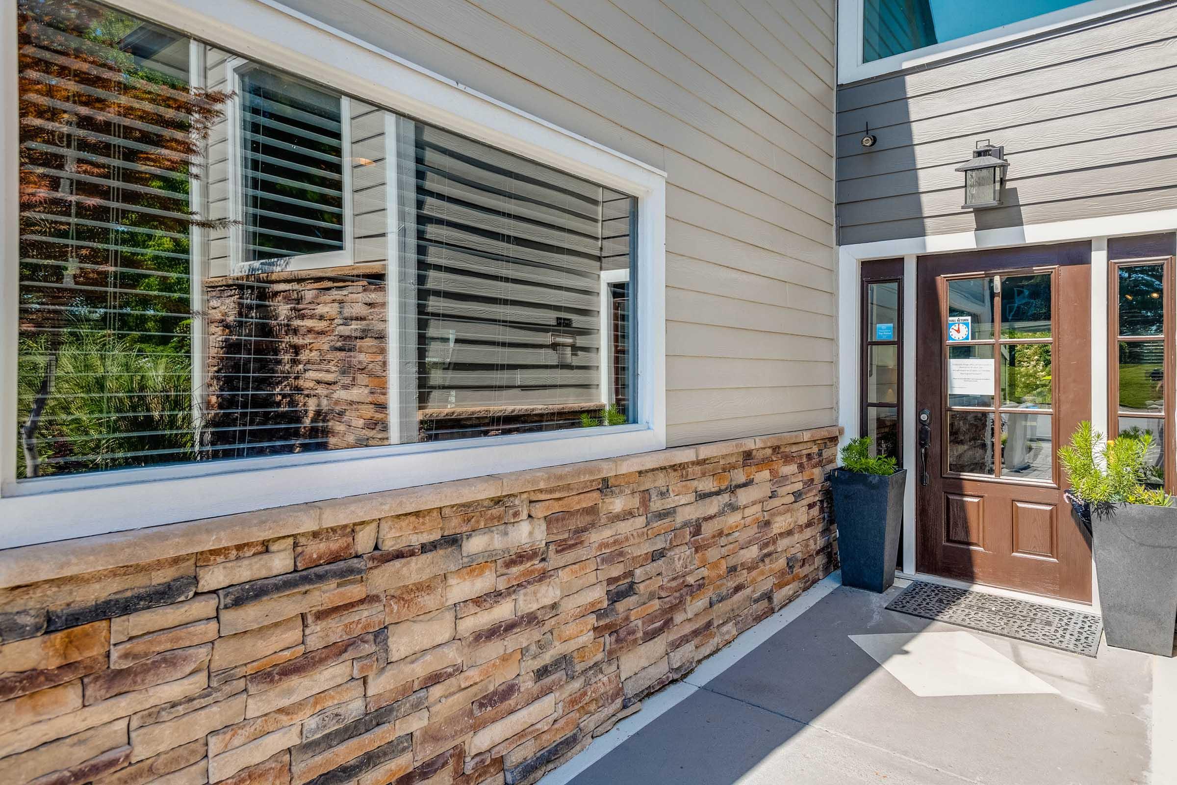 A modern building entrance featuring a wooden door with glass panes, surrounded by a stone accent wall. Large windows reflect the exterior landscape, and potted plants are positioned at the entrance, enhancing the inviting atmosphere.