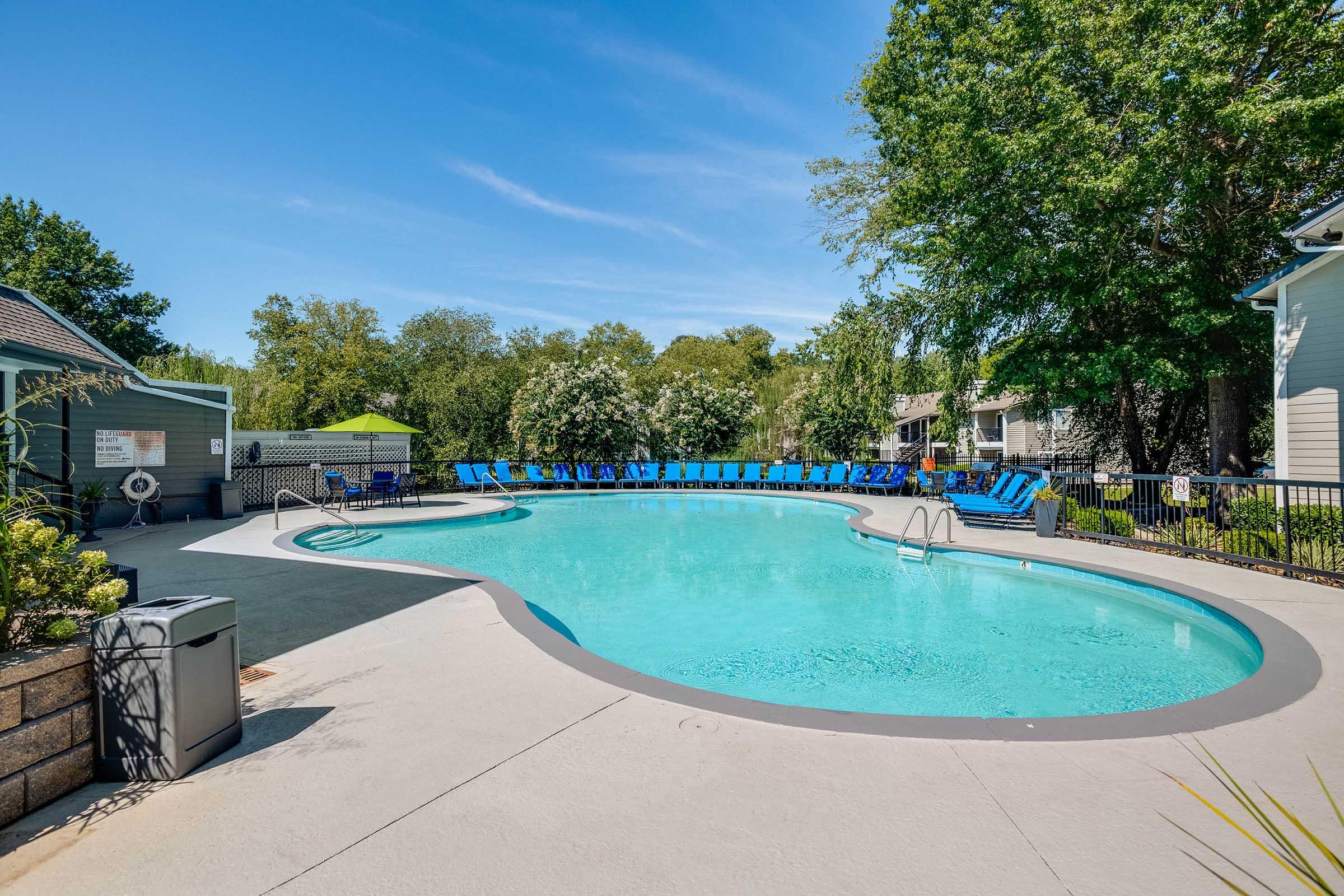 A recreational outdoor pool featuring a curved shape, surrounded by lounge chairs. Sunlight reflects off the clear water, and greenery and trees provide a natural backdrop, creating a relaxing atmosphere. A trash bin is located nearby, and there are shaded seating areas in the vicinity.
