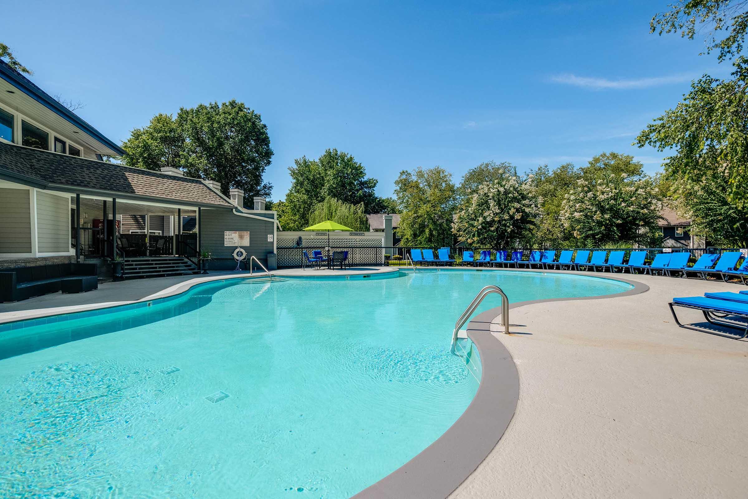 A clear, inviting swimming pool surrounded by lounge chairs. The poolside area features a smooth, light-colored deck, with greenery and trees in the background. A nearby building with large windows complements the serene outdoor setting under a blue sky.