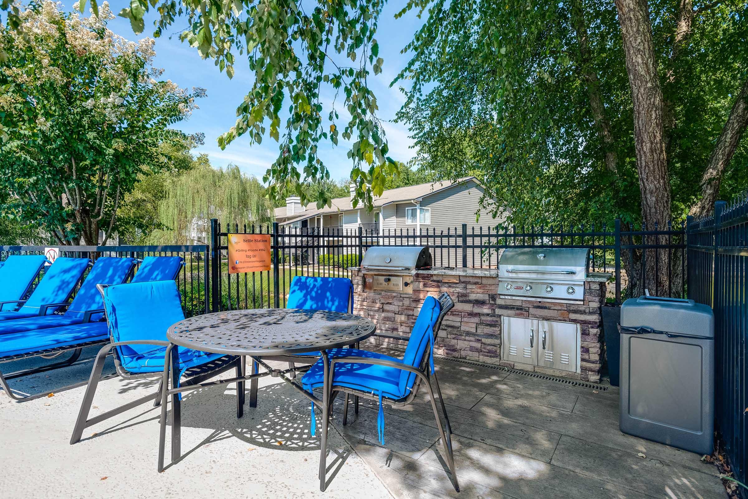A scenic outdoor area featuring a circular table with chairs, blue lounge chairs, a built-in grill station, and a trash can. Lush green trees provide shade, and a residential building is visible in the background under a clear blue sky.