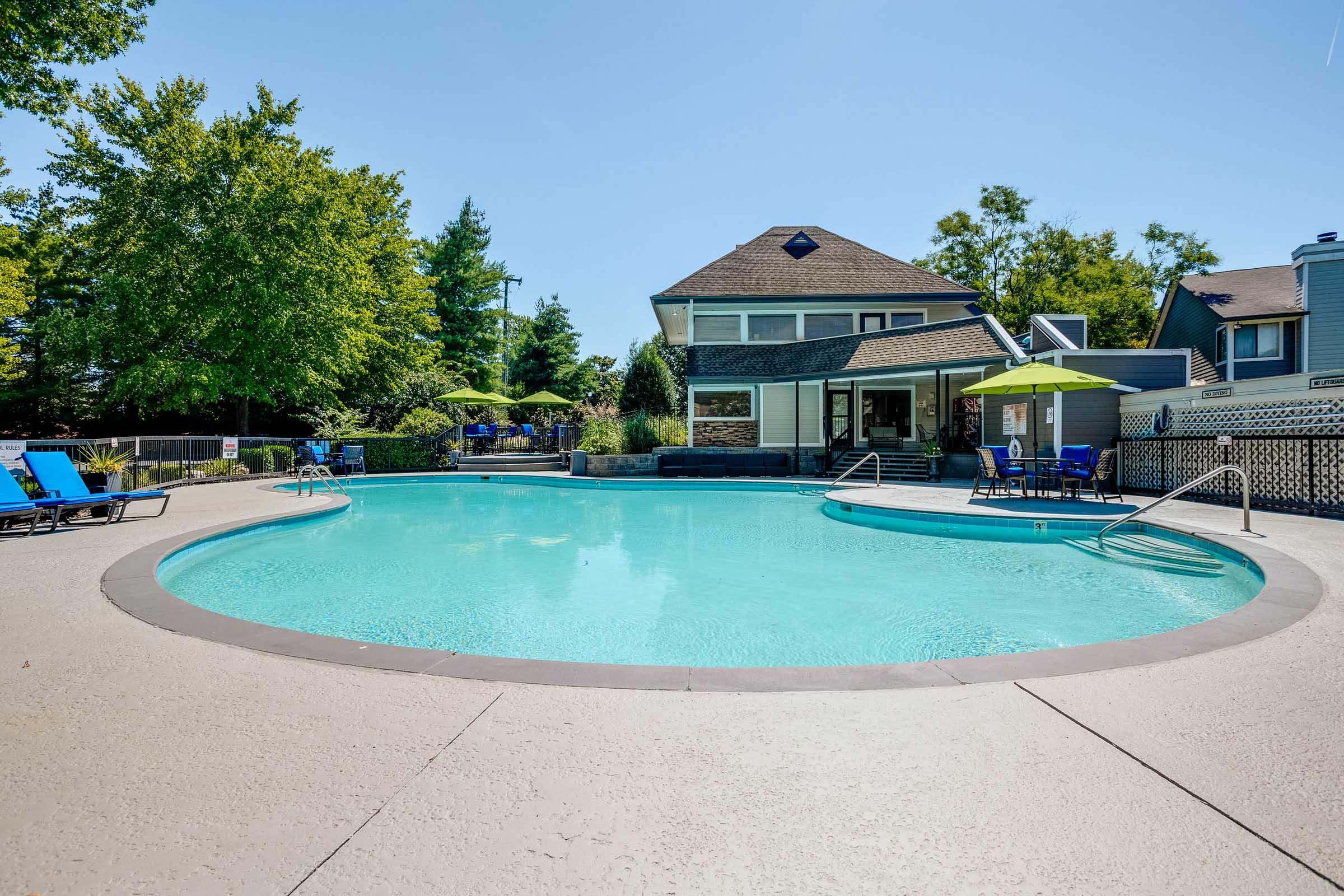 A clear blue pool surrounded by lounge chairs, with a large house and trees in the background. The sun is shining brightly in a clear sky, creating a warm and inviting outdoor atmosphere. Umbrellas provide shade near the pool area.