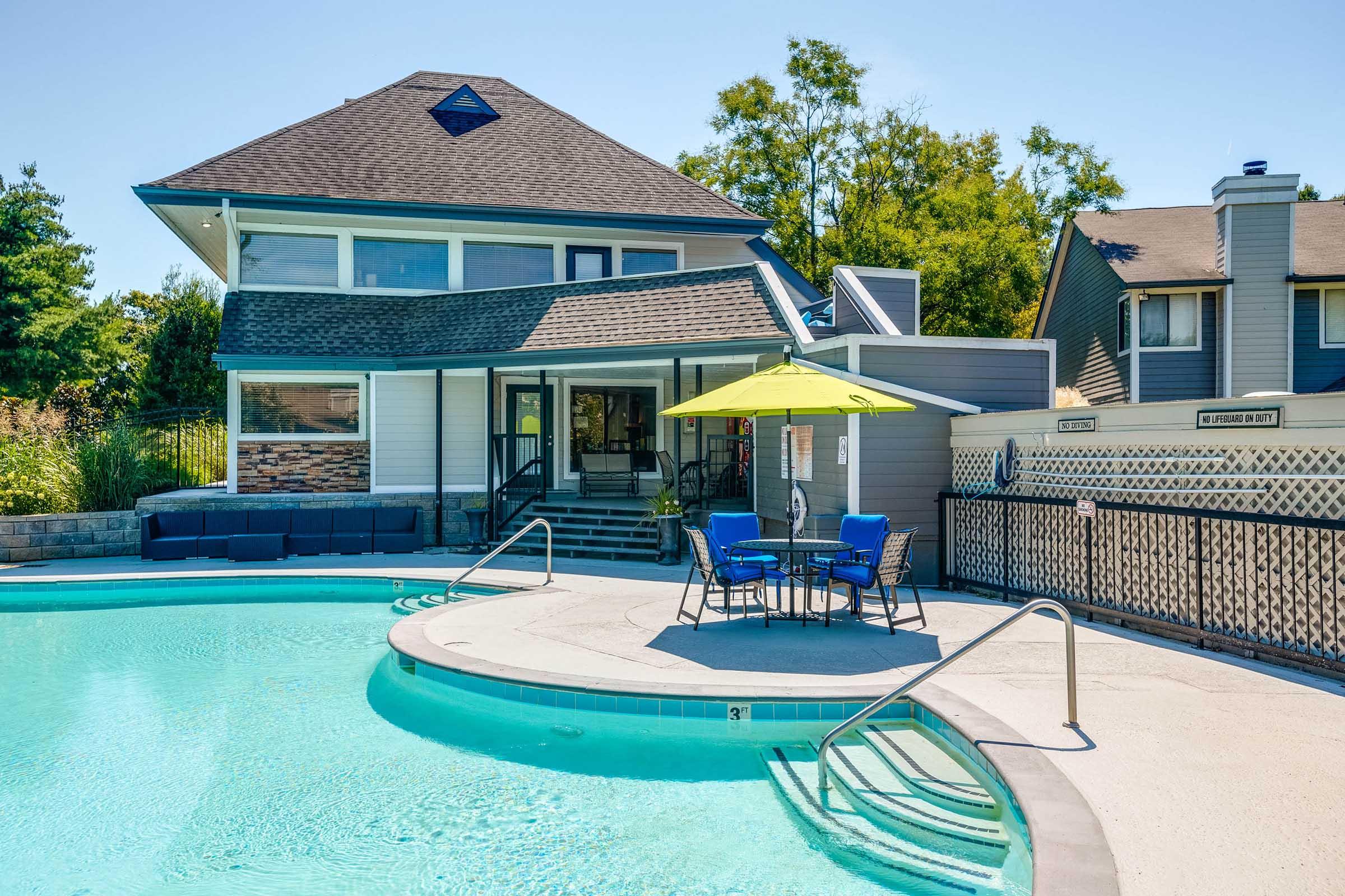 A sunny outdoor pool area featuring a curved pool with steps, a patio with a table and chairs under a green umbrella, and a modern building with large windows and a sloped roof in the background. Lush greenery surrounds the space, creating a relaxing atmosphere.