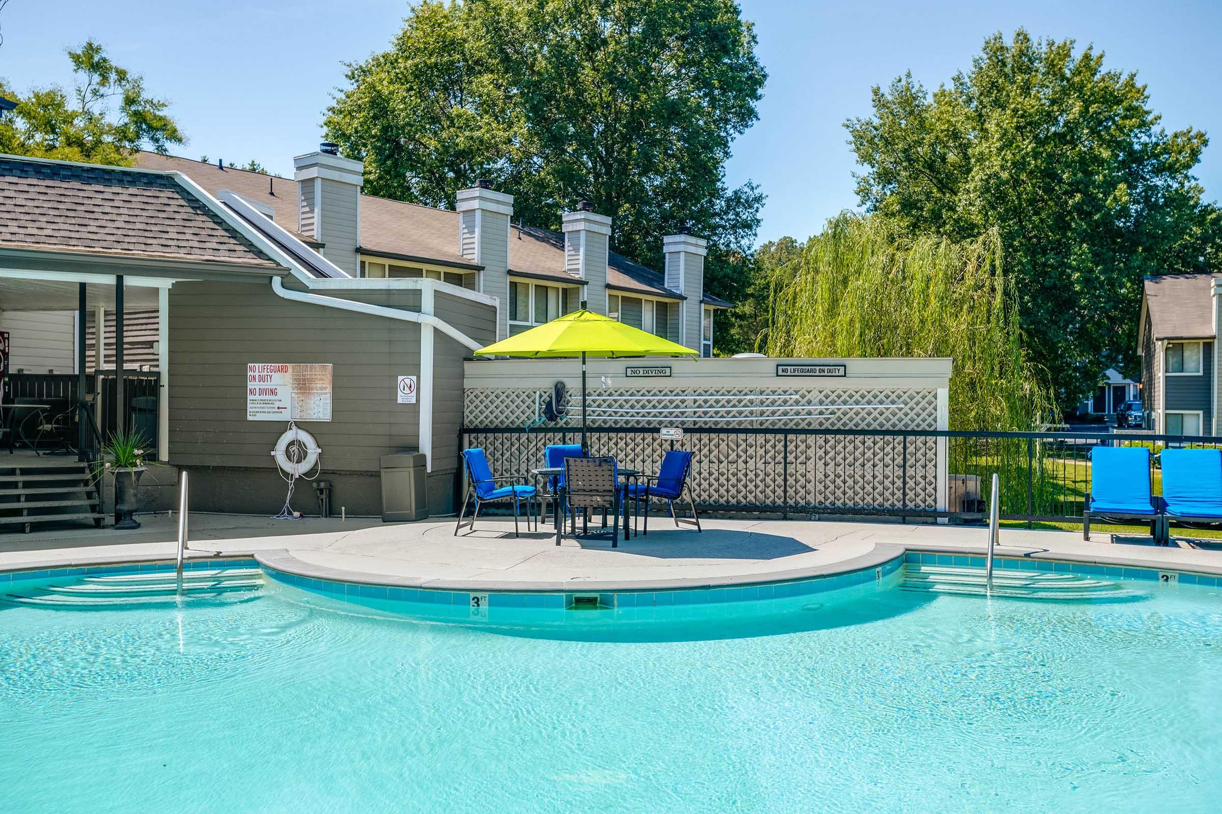 A swimming pool area featuring a circular pool with clear blue water, surrounded by lounge chairs and a small table under a bright green umbrella. In the background, there are several buildings with trees providing shade. The scene reflects a sunny day, perfect for relaxation.
