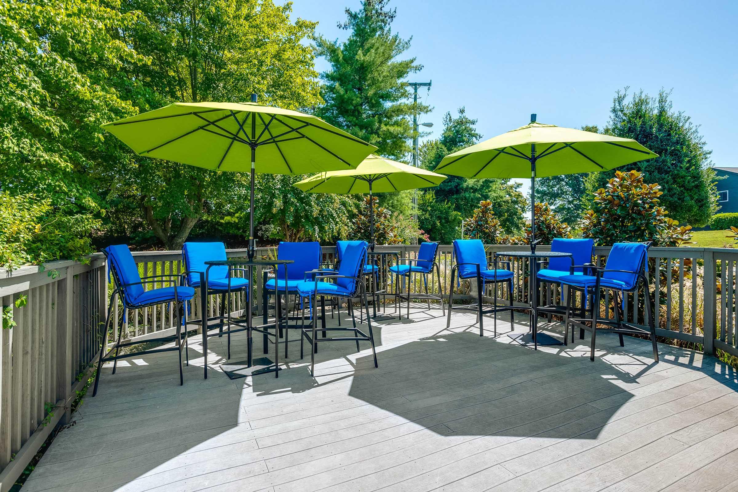 A spacious outdoor deck featuring several high-top tables with bright blue chairs and large green umbrellas, surrounded by lush greenery and trees under a clear blue sky.